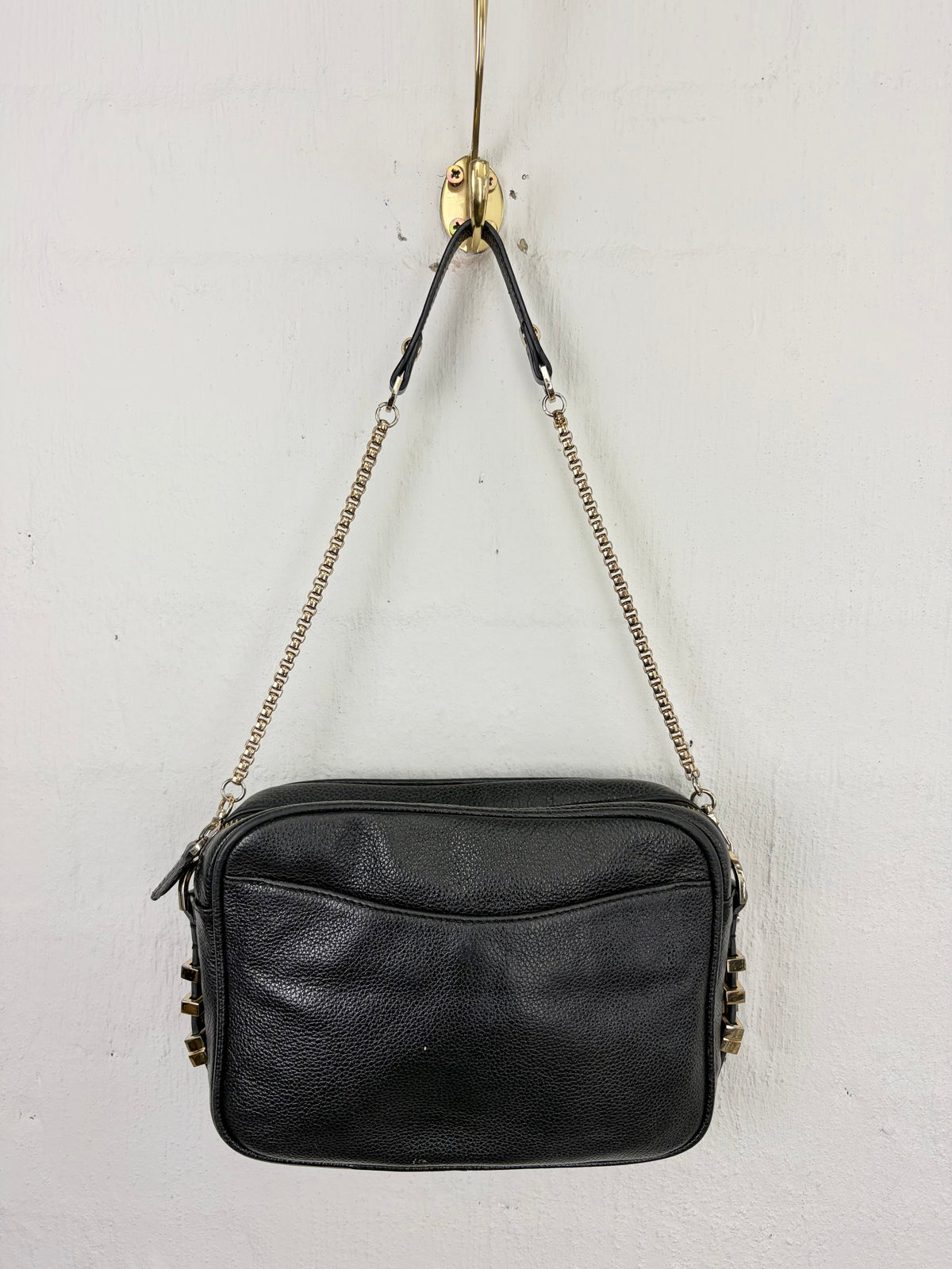 A black leather crossbody camera bag with a gold-tone chain strap, hanging from a gold wall hook against a white textured wall.