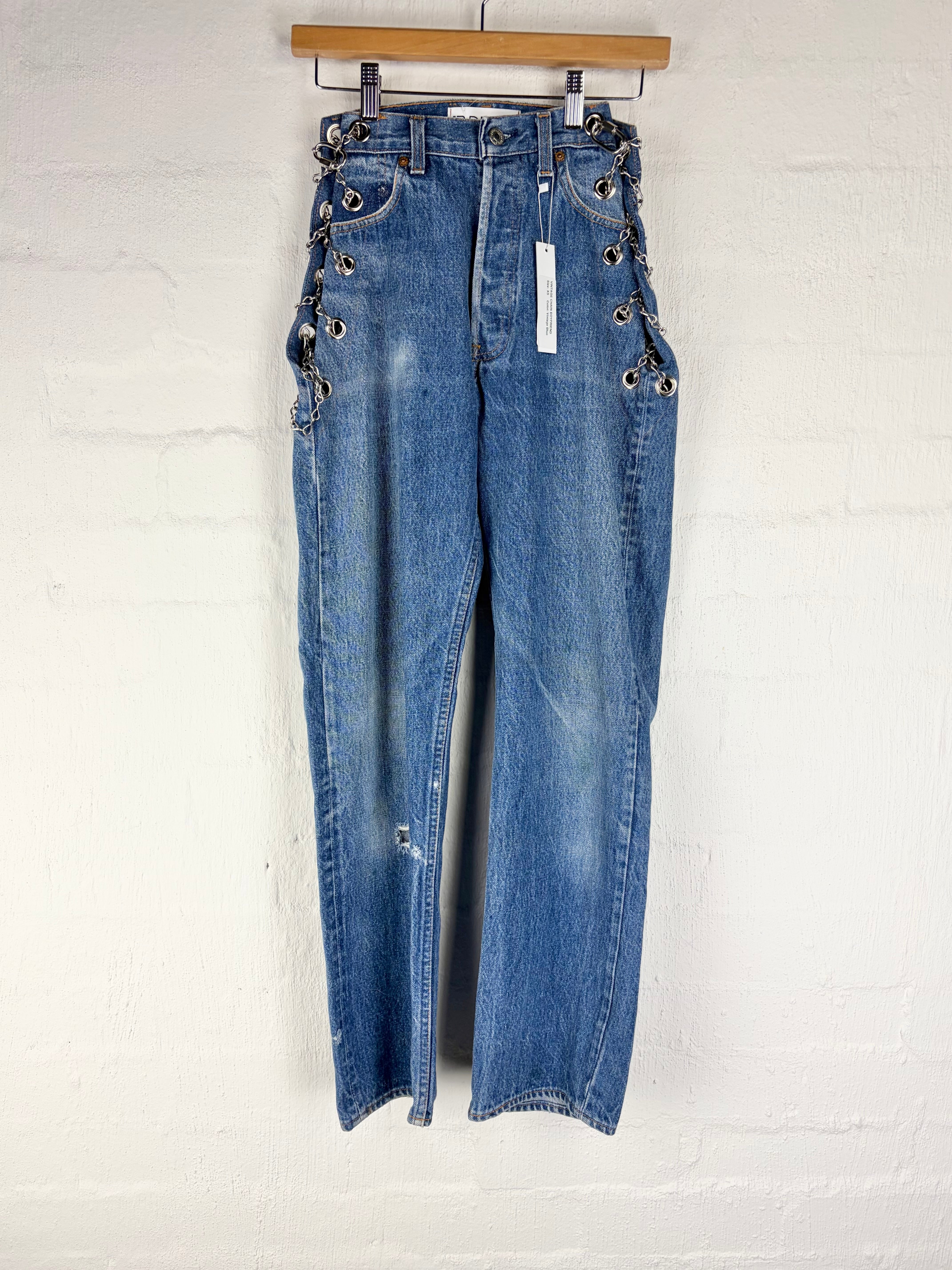 A pair of high-waisted blue denim jeans with decorative metal eyelets and lace-up detailing on the sides, hanging from a wooden hanger against a white painted brick wall.