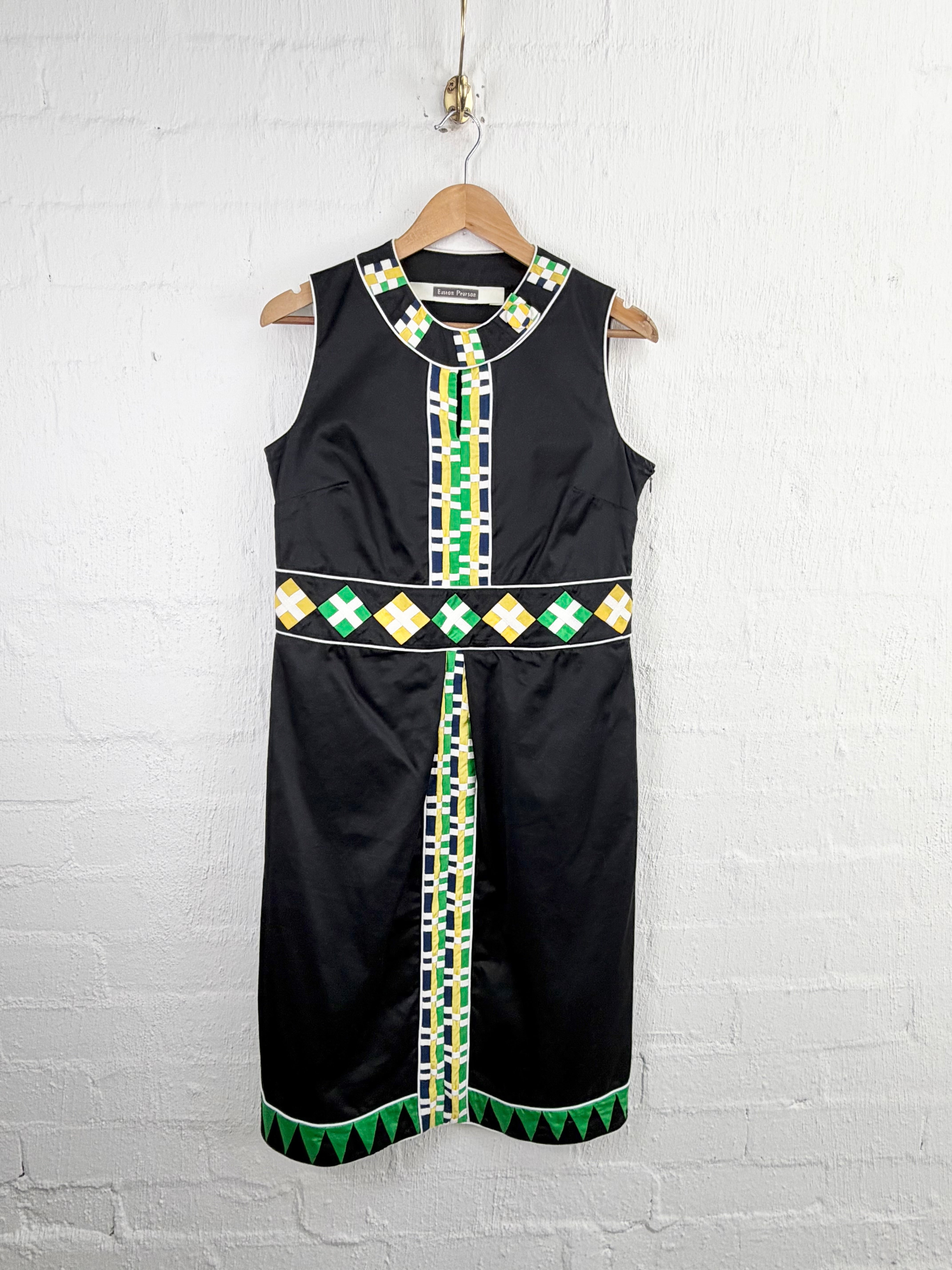 A sleeveless black dress with green, white, and yellow geometric patterns, hanging on a wooden hanger against a white brick wall.