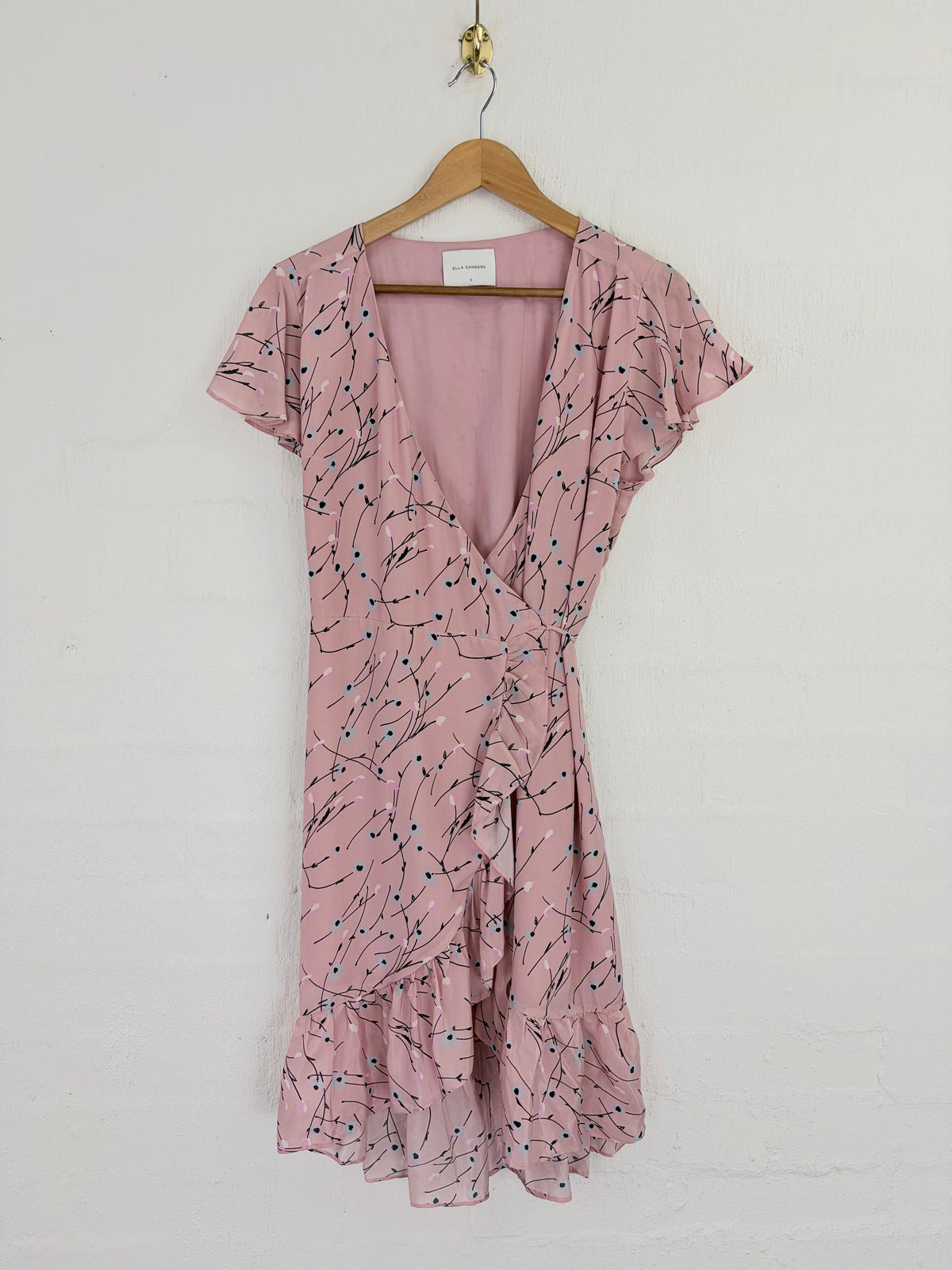 A pink short-sleeve wrap dress with a dark blue and black abstract line print, hanging on a wooden hanger against a white painted brick wall.