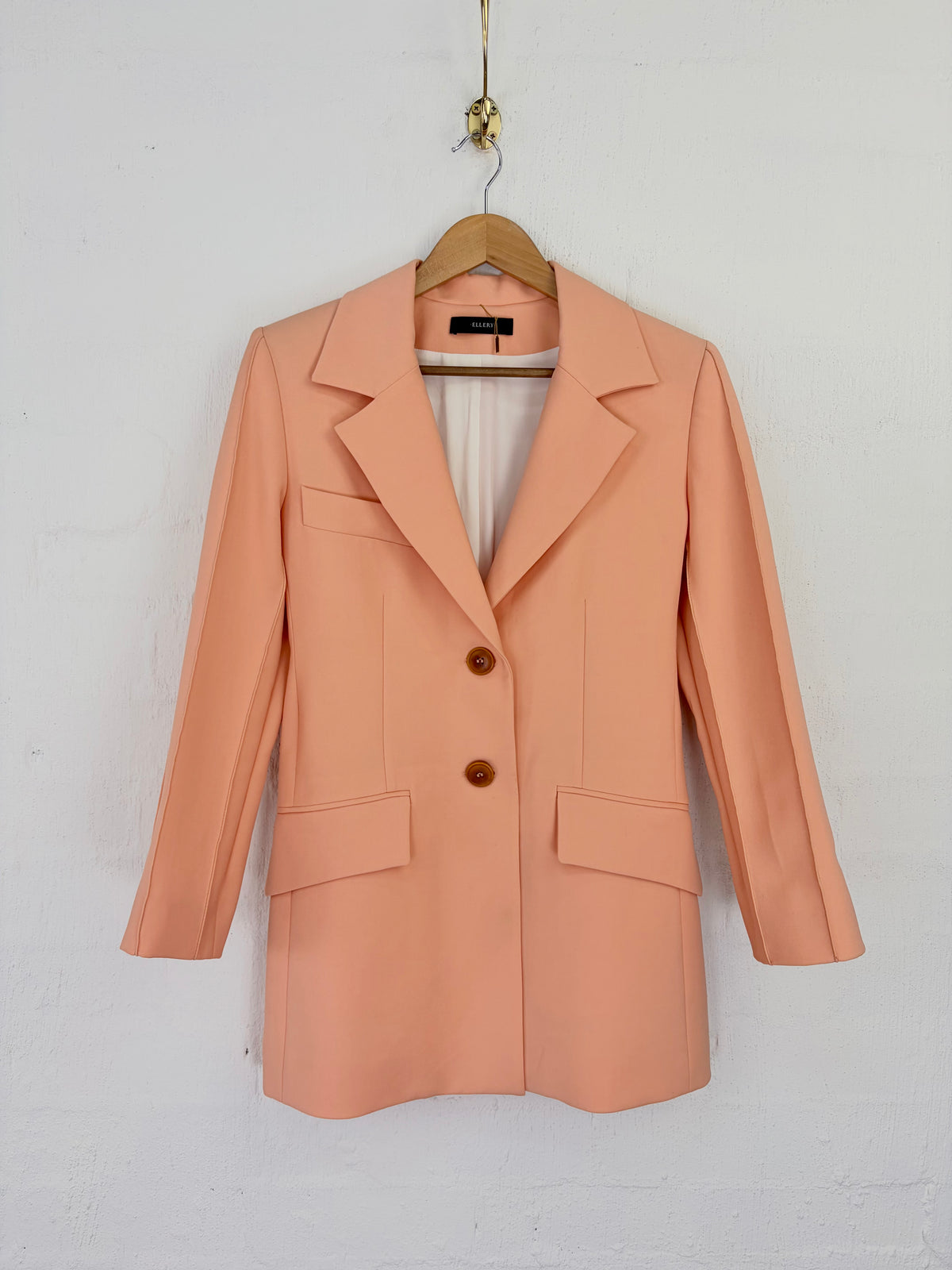 A peach-colored double-breasted blazer with notched lapels and two front buttons, hanging on a wooden hanger against a white painted brick wall.