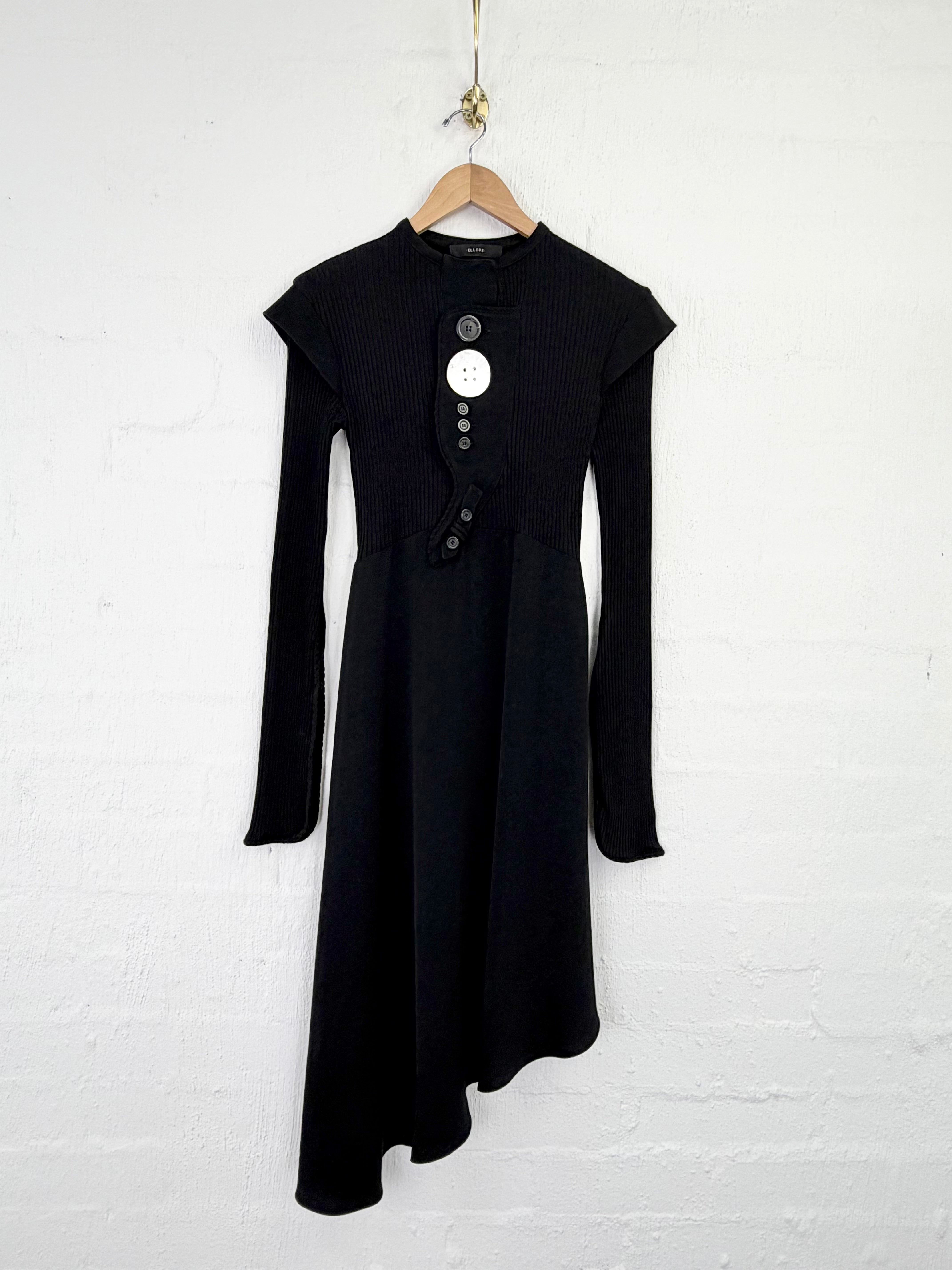 A black long-sleeve midi dress with a high-low asymmetrical hem and a buttoned neckline, hanging on a wooden hanger against a white painted brick wall.