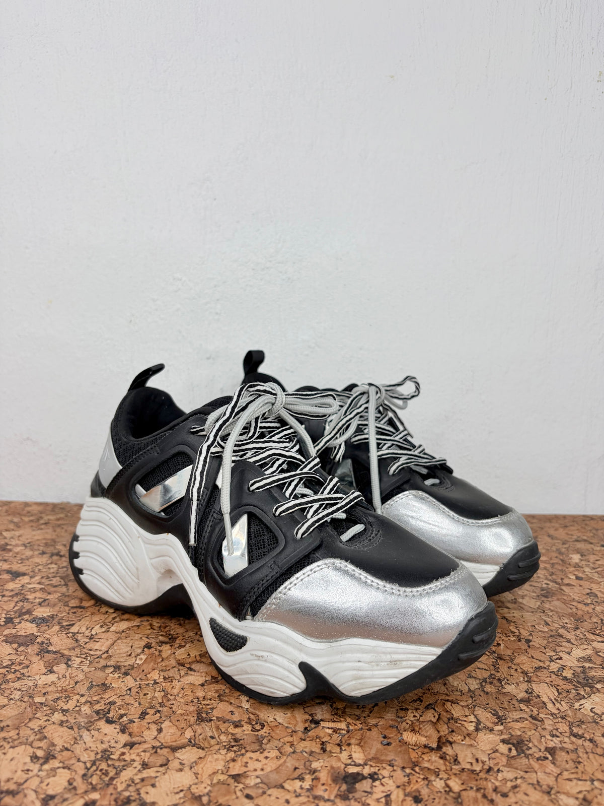 A pair of chunky low-top sneakers in black and metallic silver, featuring white laces and a thick sculpted white and black sole, displayed on a cork surface against a plain wall.