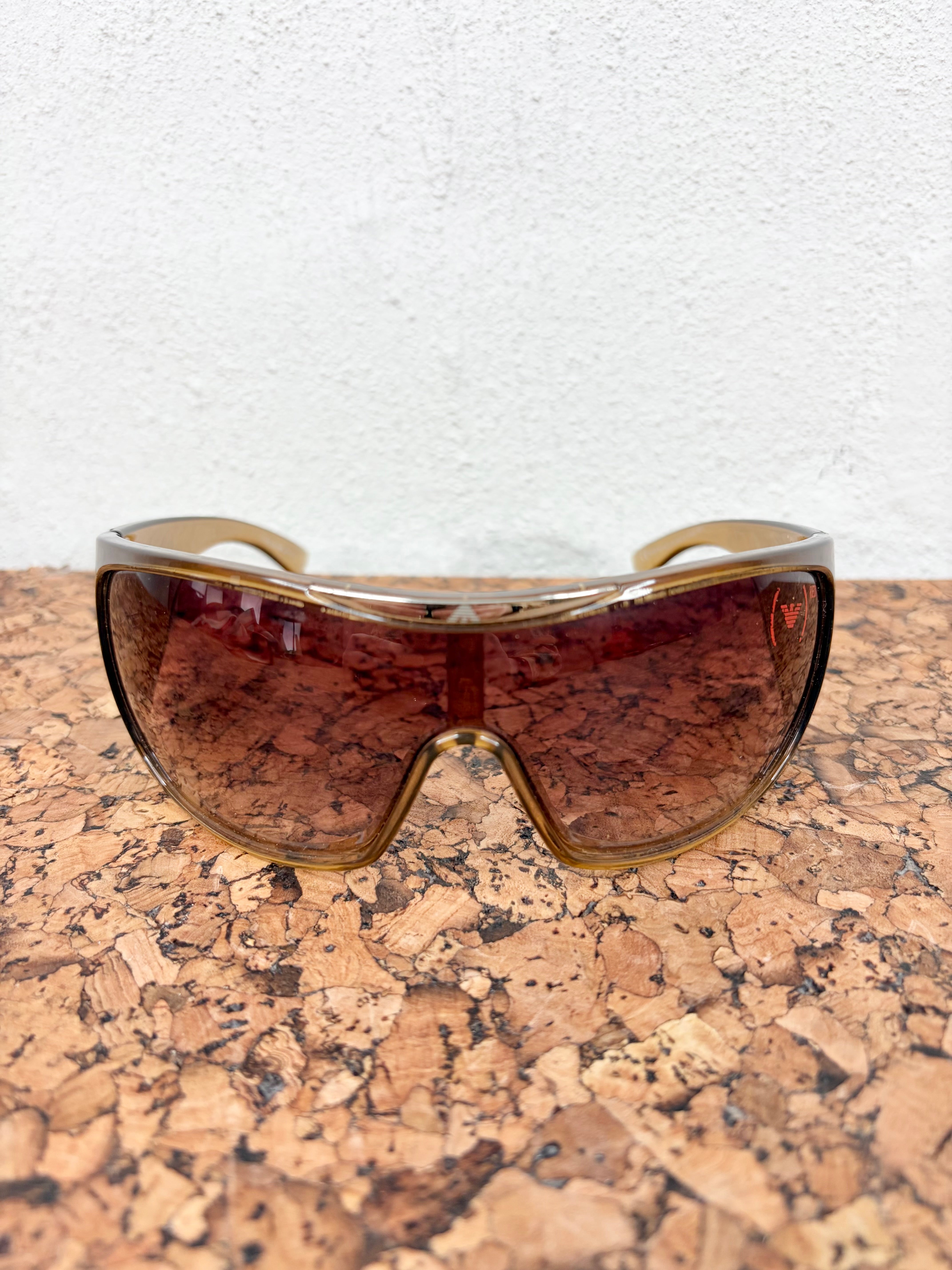 Shield-style safety sunglasses with a translucent amber-tinted frame and dark brown mirrored lenses, resting on a cork surface against a white wall.