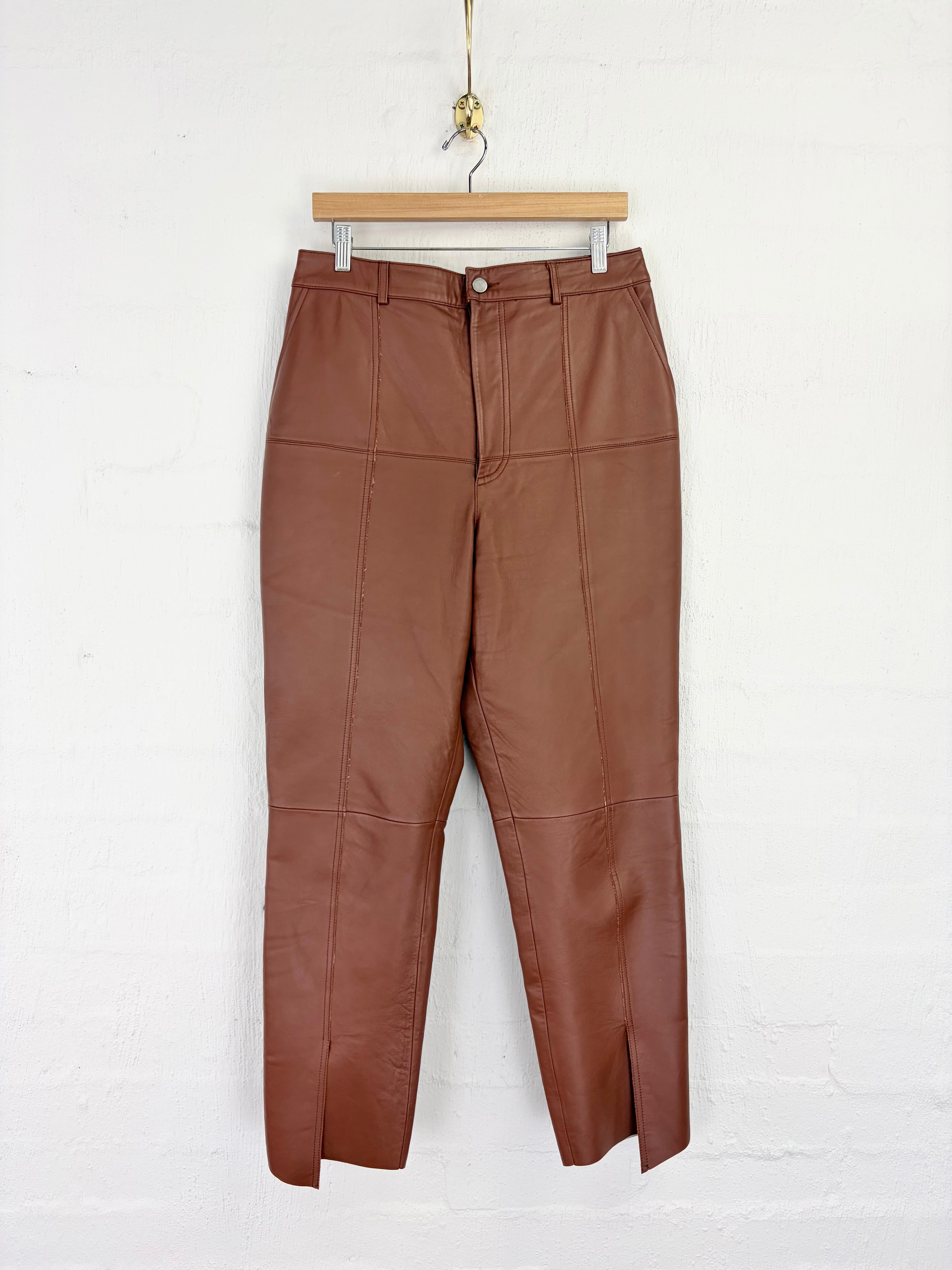 A pair of brown faux leather cropped pants with a patchwork panel design, hanging from a wooden hanger against a white brick wall.