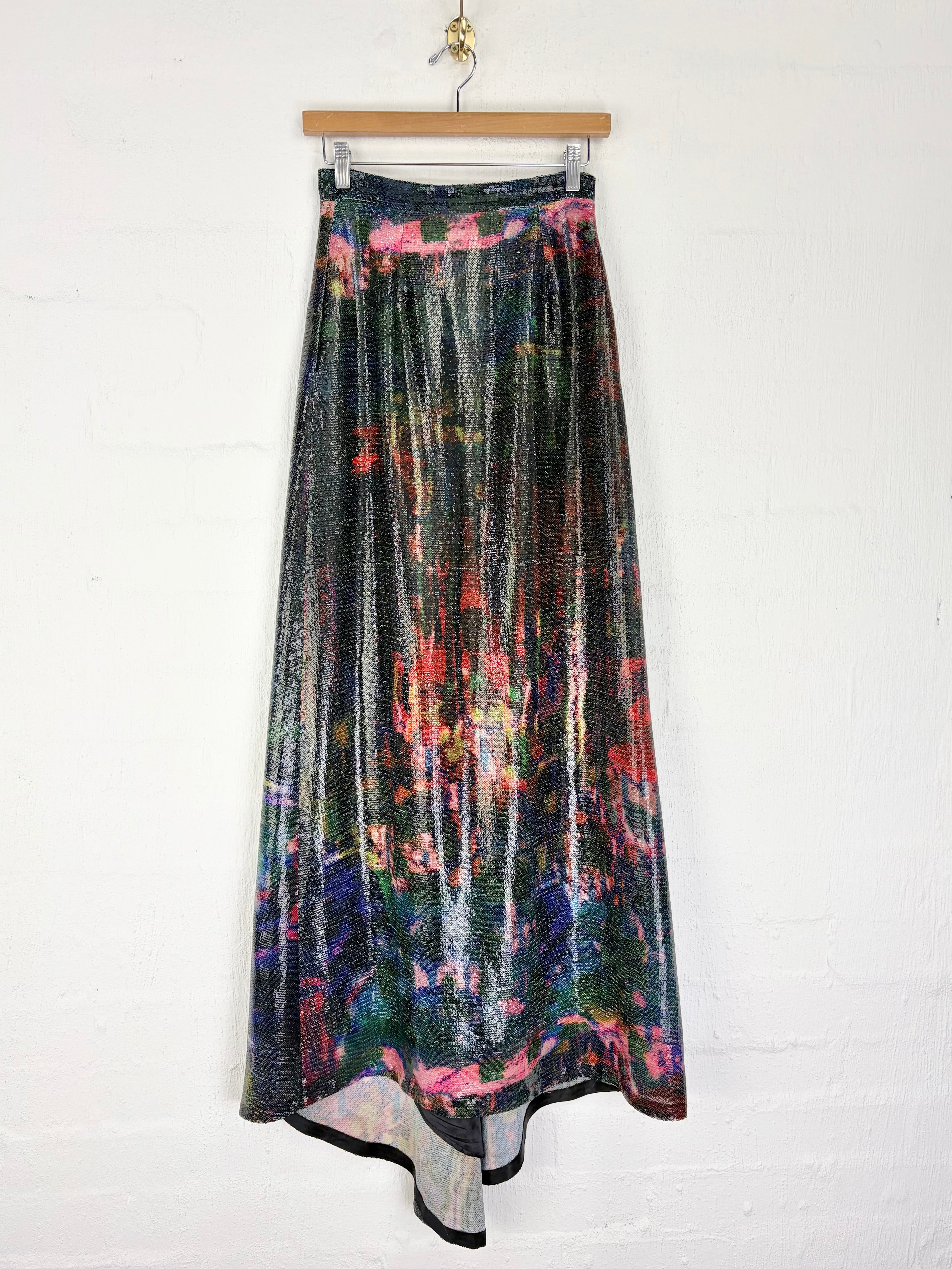 A high-waisted, floor-length skirt with a metallic, crinkled texture and an abstract multicolor print, hanging from a wooden hanger against a white painted brick wall.
