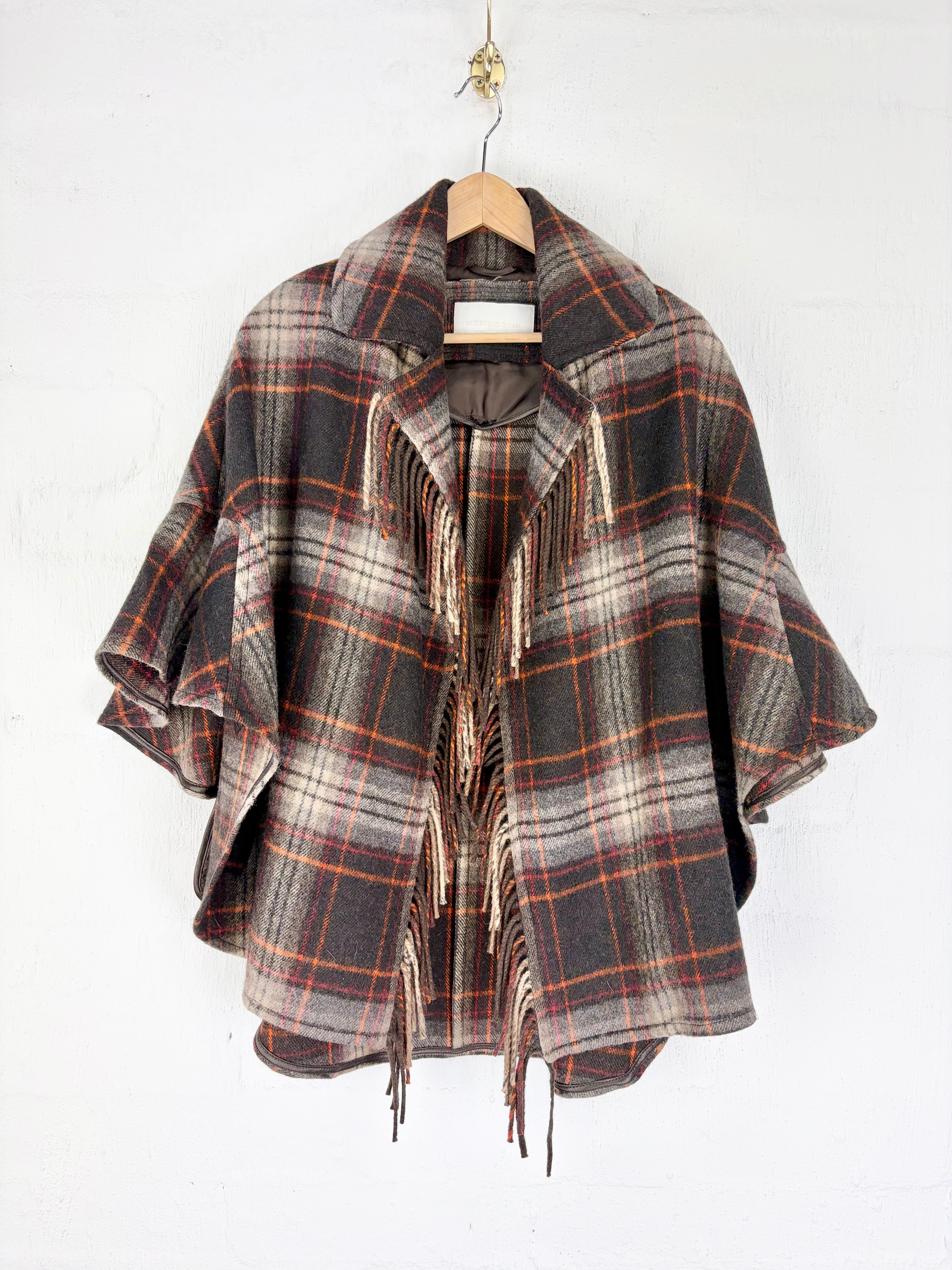 A plaid fringed cape jacket in a brown, gray, and orange tartan pattern, hanging on a wooden hanger against a white wall.