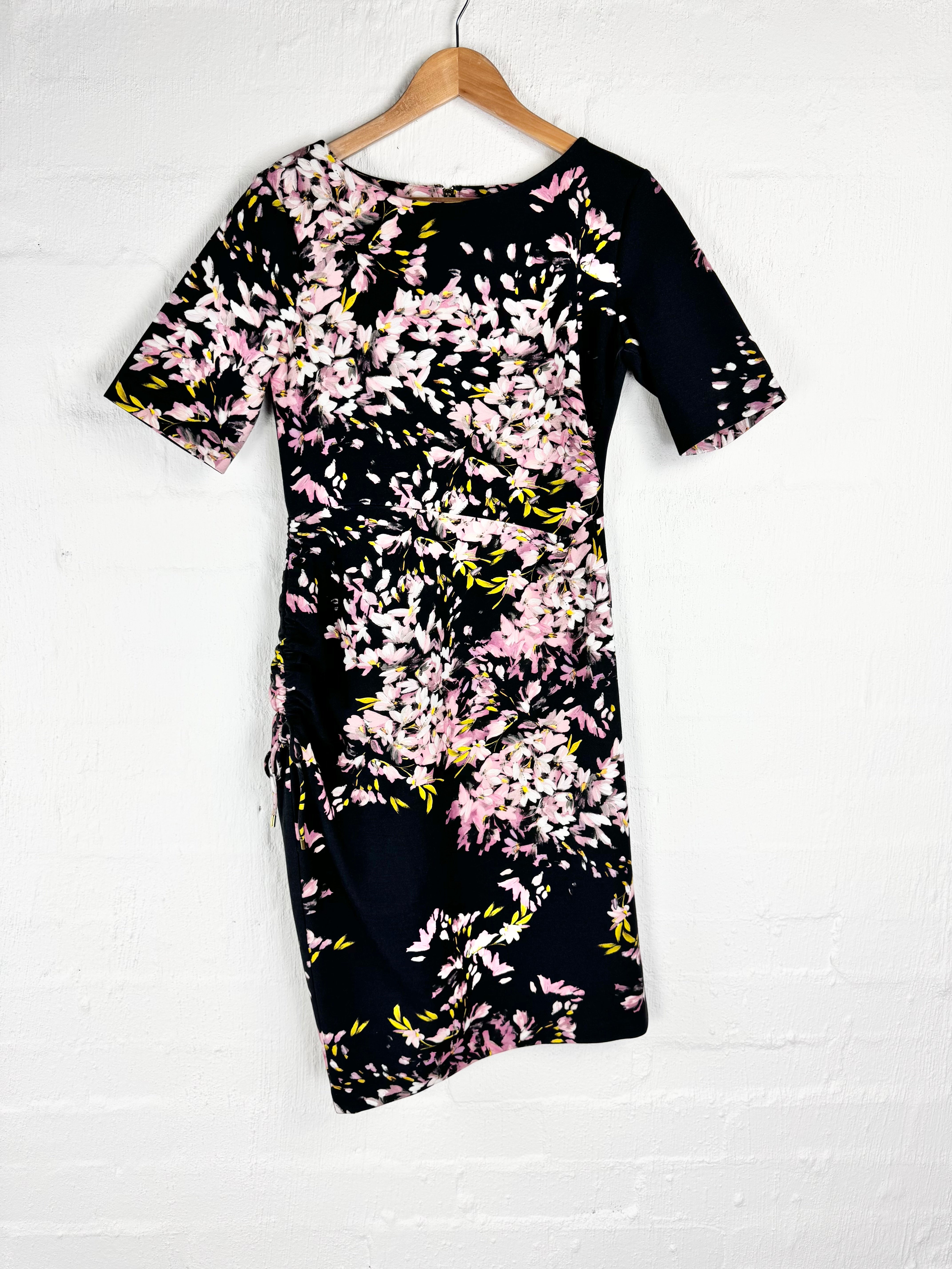 A short-sleeve bodycon dress with a black base and a pink, white, and yellow floral print, hanging on a wooden hanger against a white brick wall.