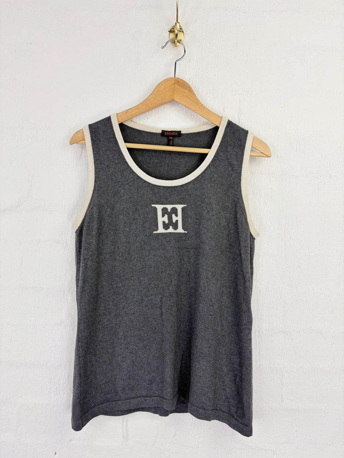 A dark gray heathered tank top with cream-colored trim and a white embroidered Tommy Hilfiger logo on the chest, hanging on a wooden hanger against a white brick wall.