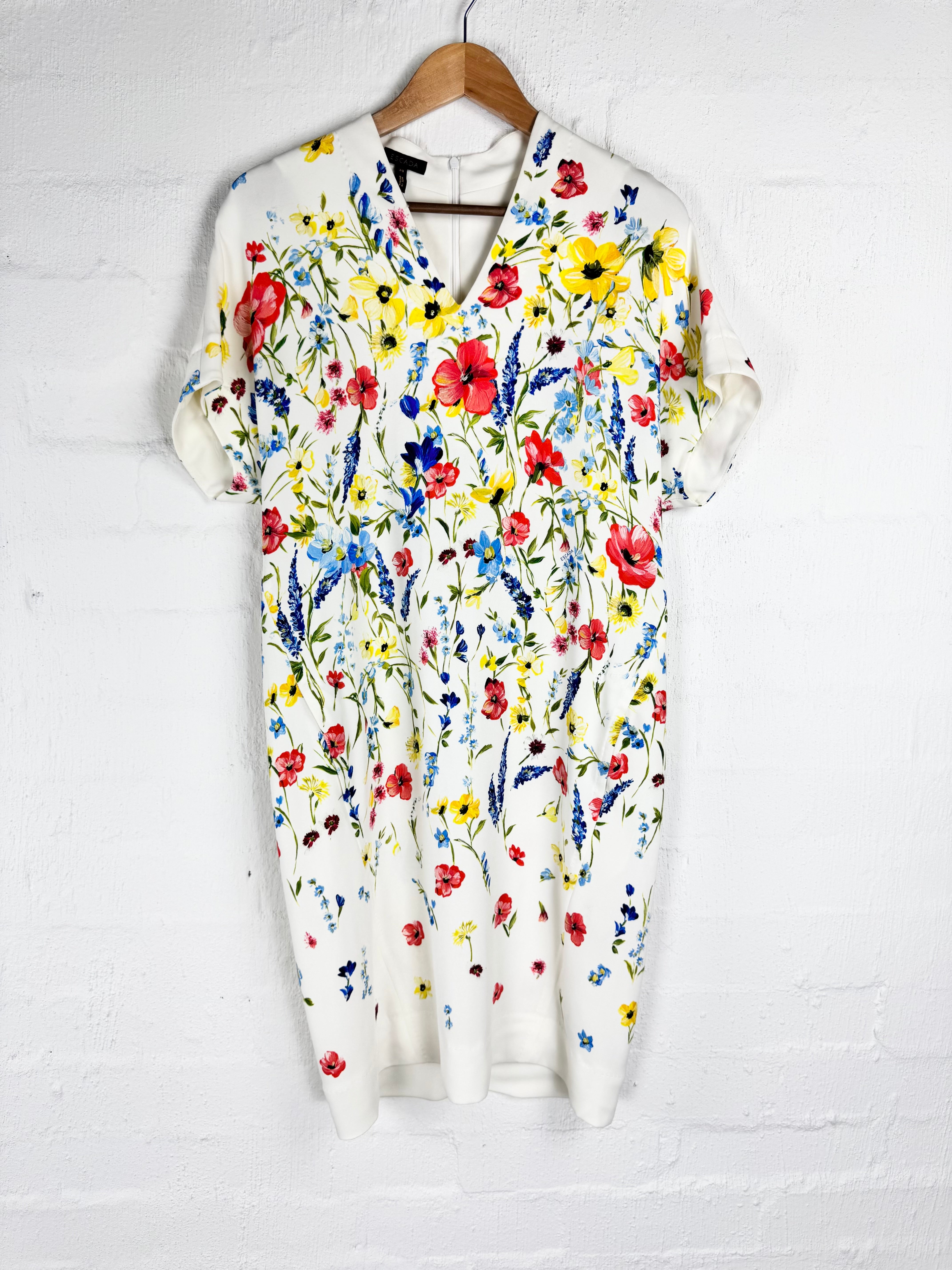 A short-sleeve V-neck knit dress with an all-over colorful wildflower print, hanging on a wooden hanger against a white painted brick wall.
