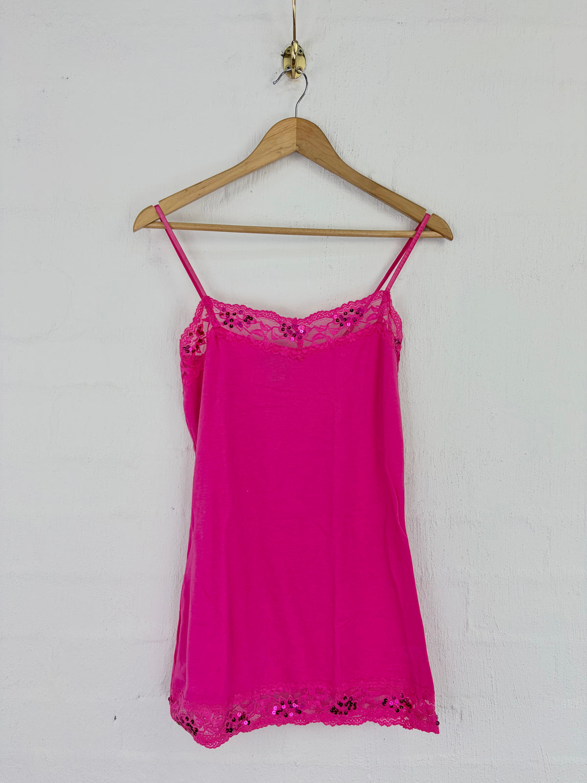 A bright pink camisole top with thin straps and floral lace trim at the neckline and hem, hanging on a wooden hanger against a white wall.