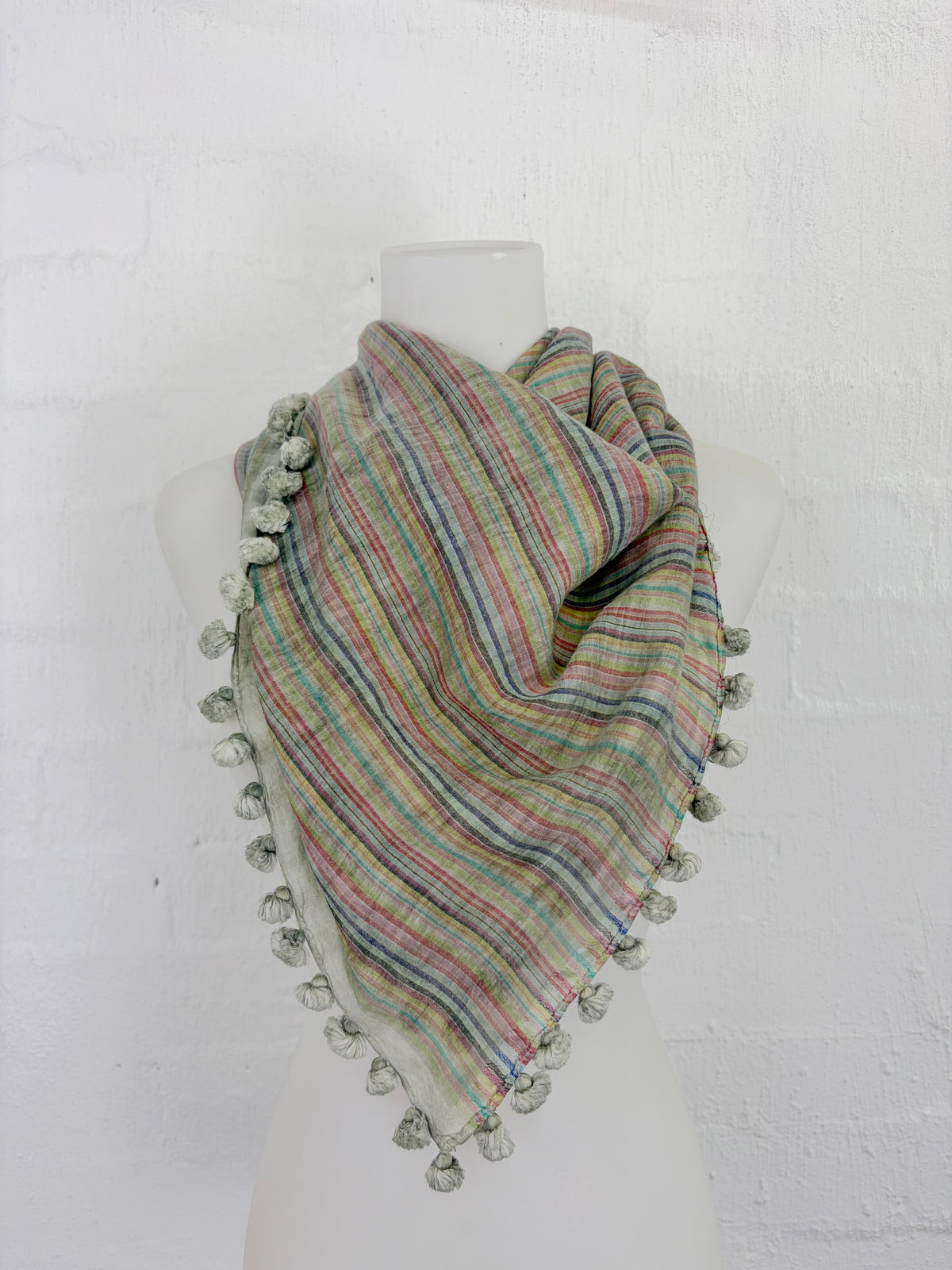 A triangular striped scarf with pom-pom trim draped on a white mannequin torso against a white brick wall background.