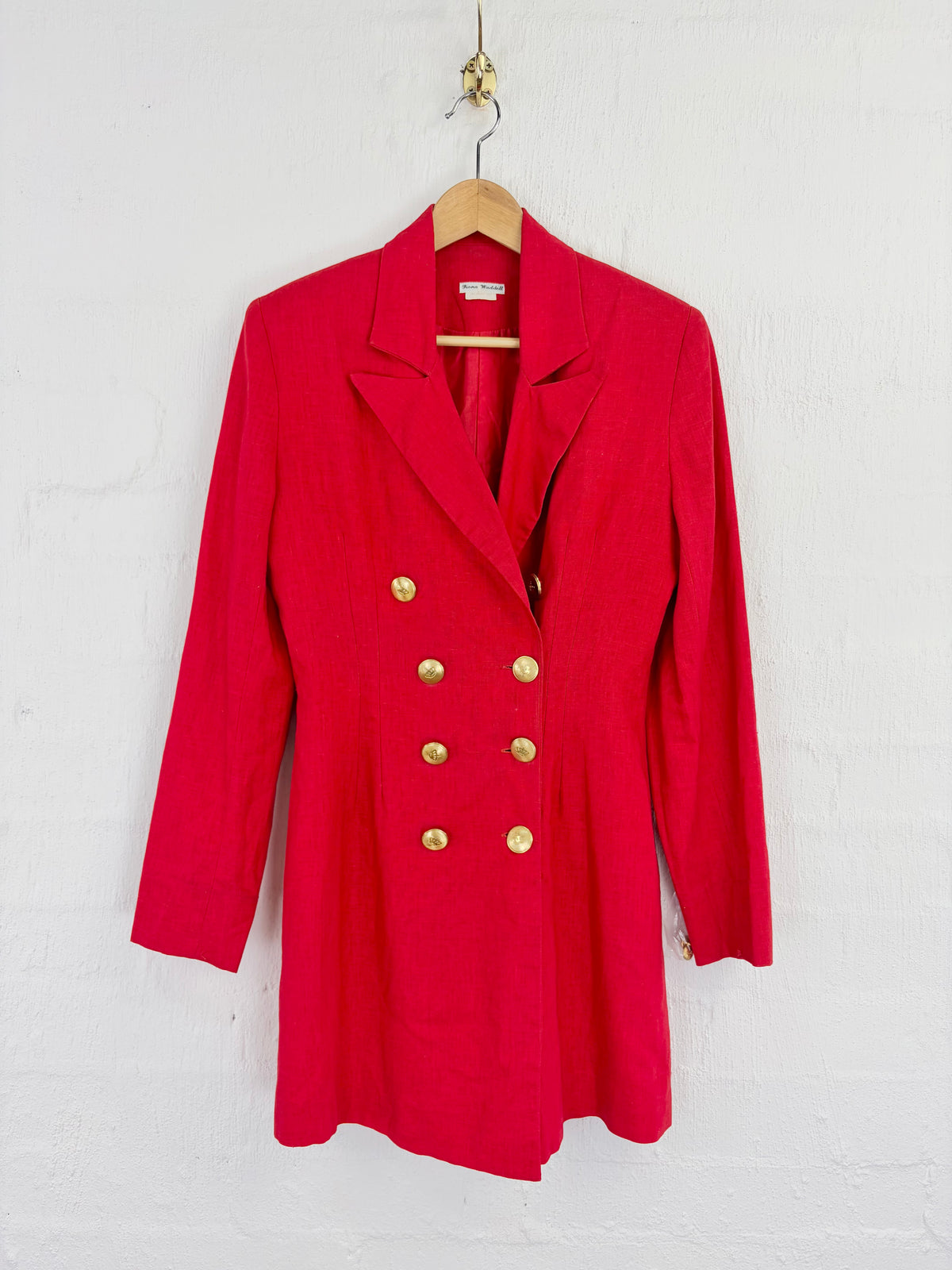A bright red double-breasted blazer with gold buttons, hanging on a wooden hanger against a white painted brick wall.