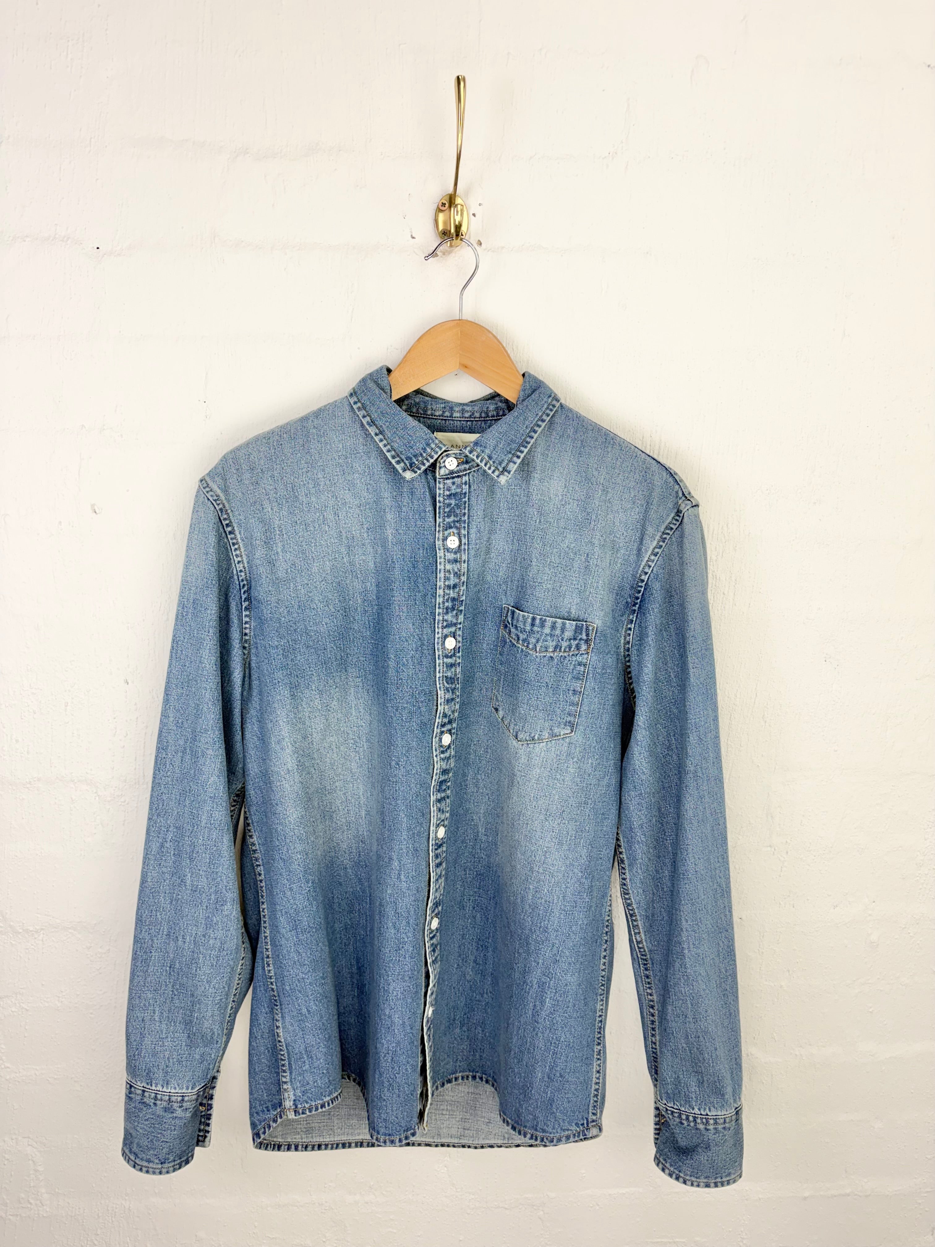 A light blue denim long-sleeve button-up shirt hanging on a wooden hanger against a white painted brick wall.