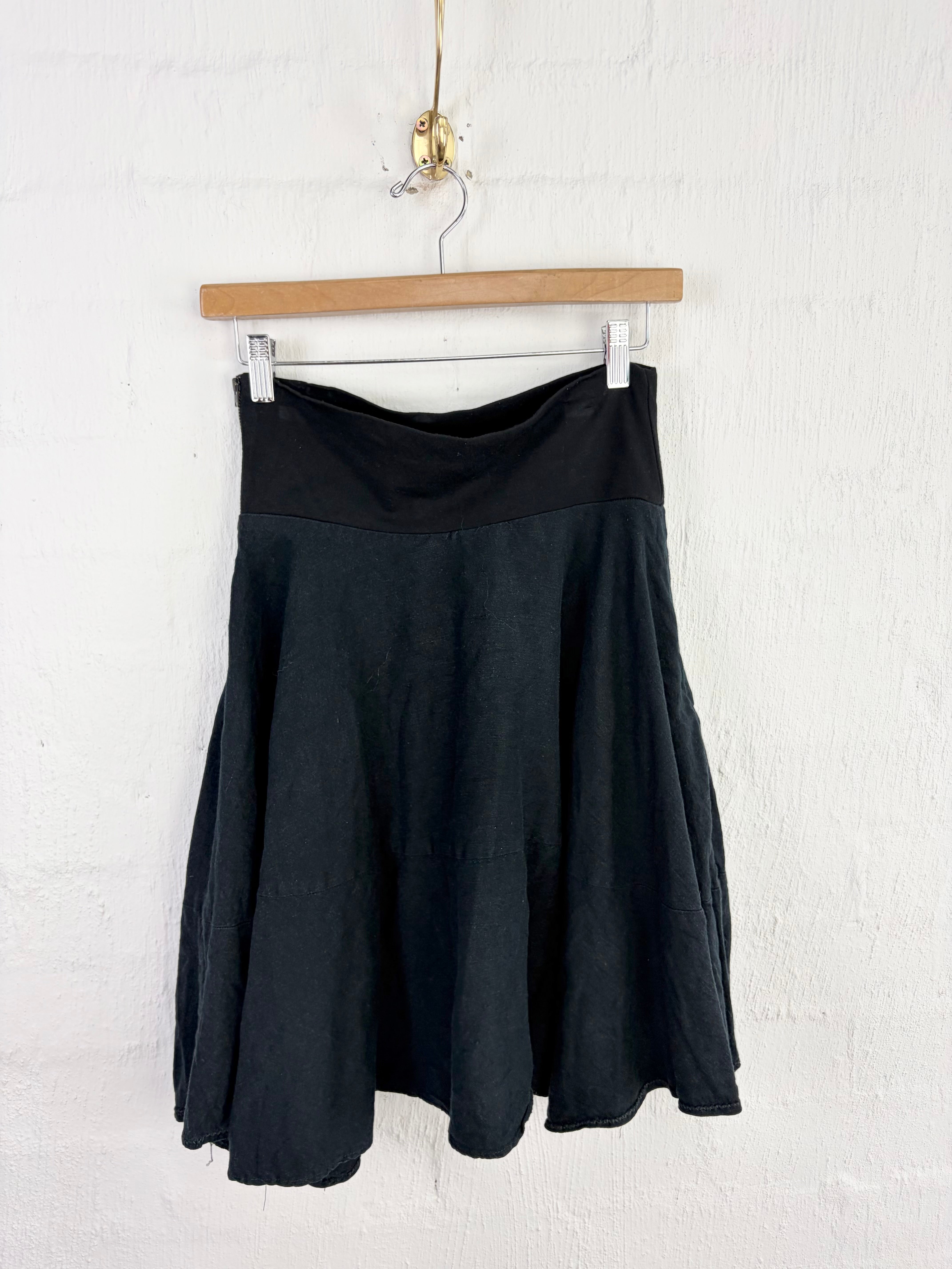 A black flared skirt with a wide black waistband, hanging from a wooden hanger against a white painted brick wall.