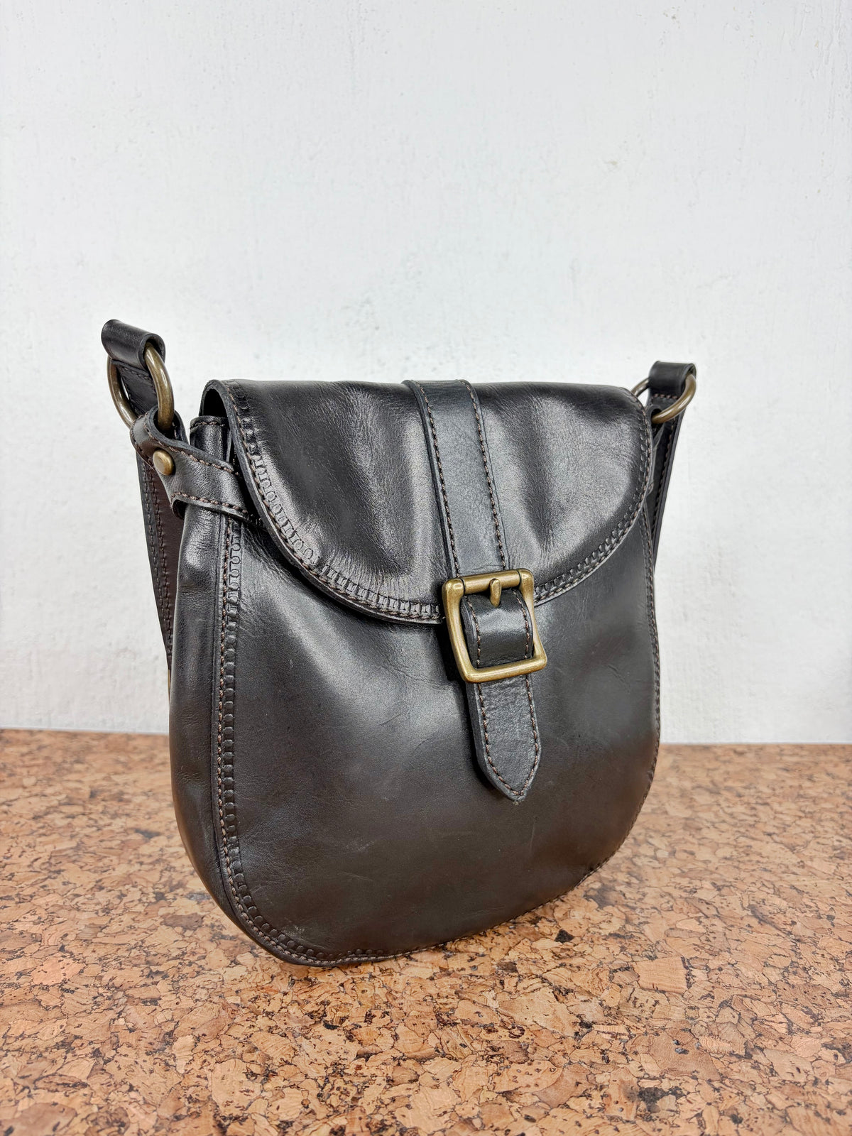 A dark brown leather shoulder bag with a flap closure and a brass buckle, resting on a cork surface against a white wall.