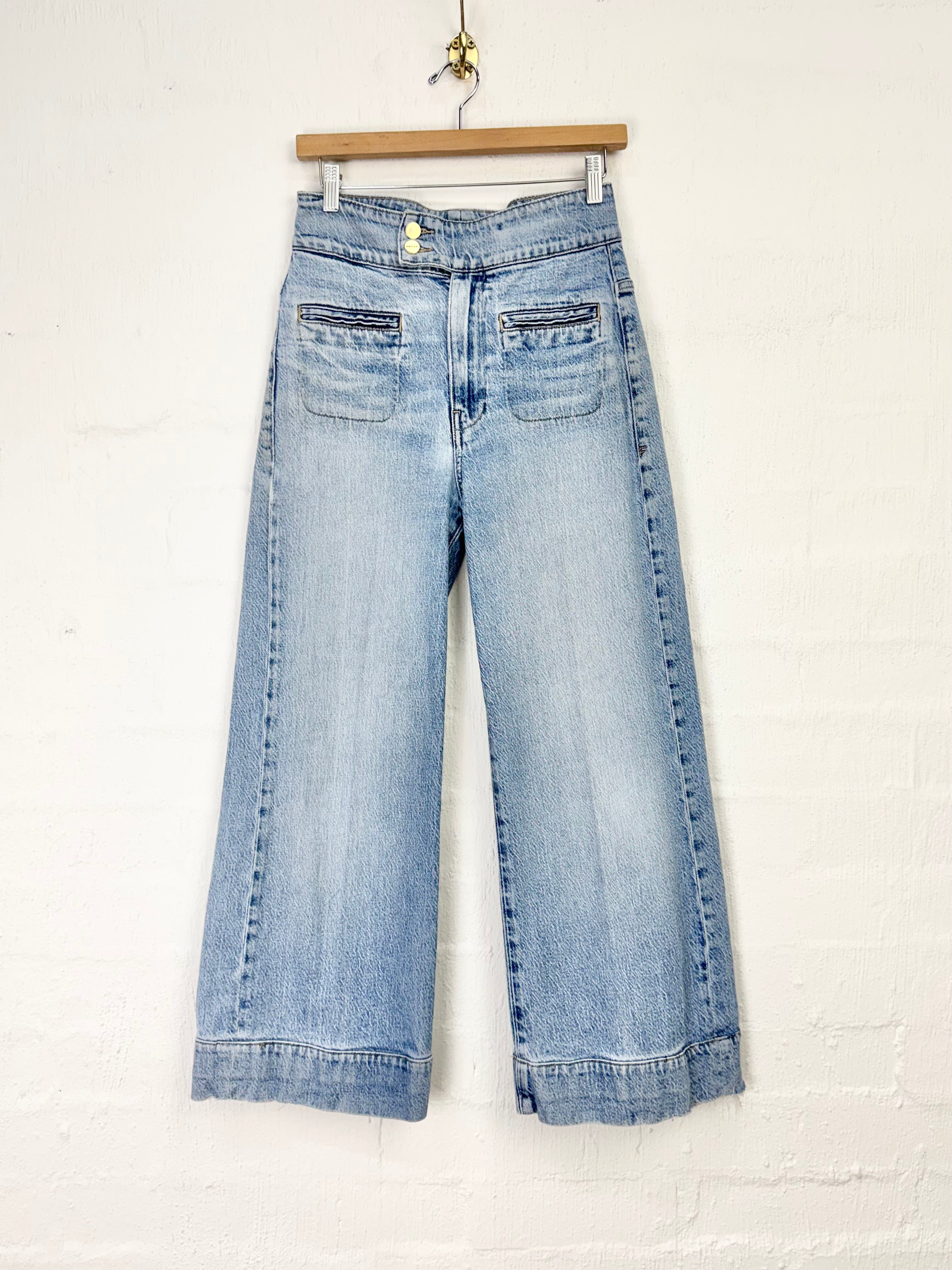 Light blue wide-leg denim jeans hanging from a wooden hanger against a white painted brick wall.