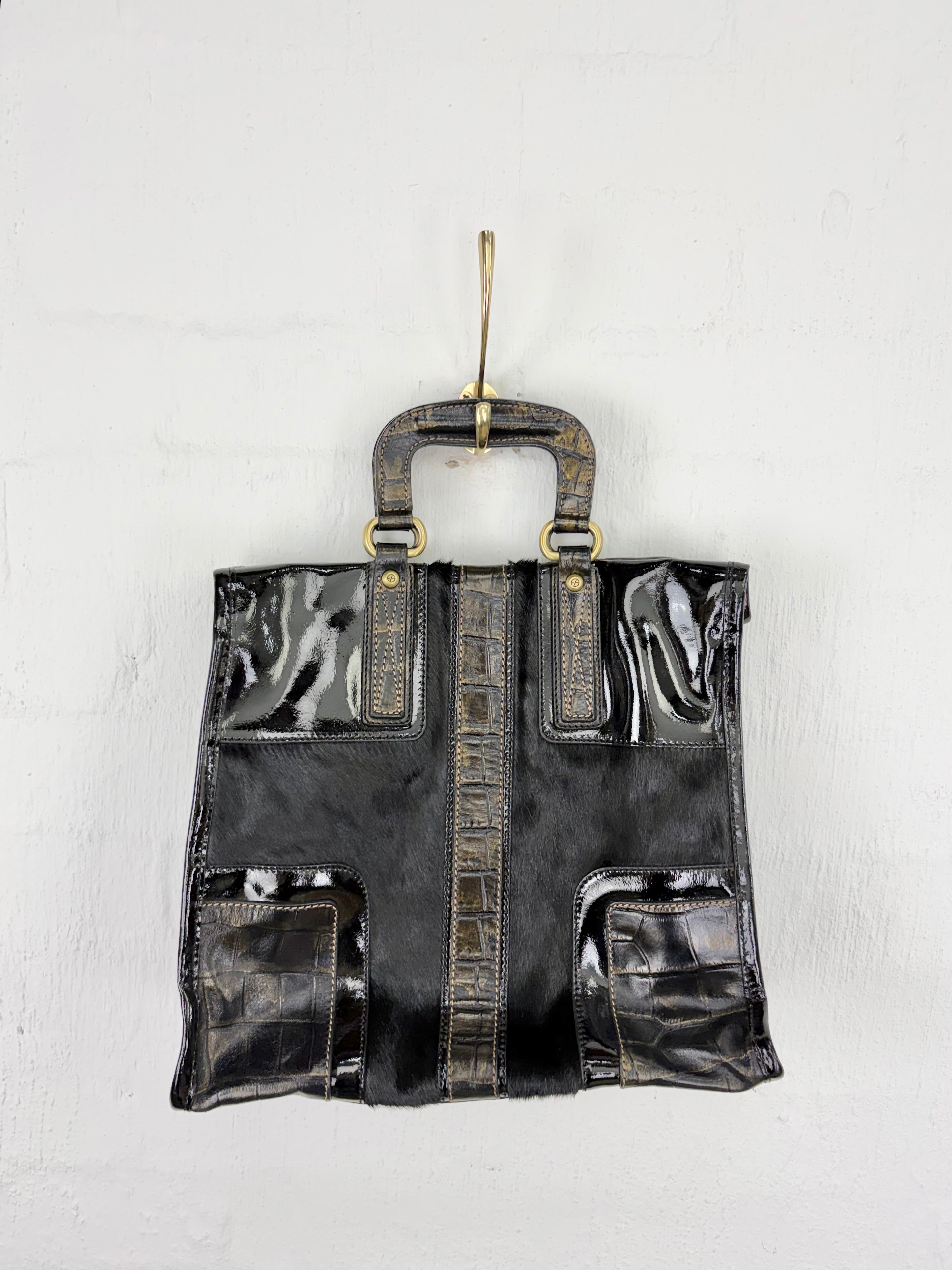 A black handbag with glossy patent leather panels, textured faux-croc details, and a top handle, hanging from a gold hook against a white wall.
