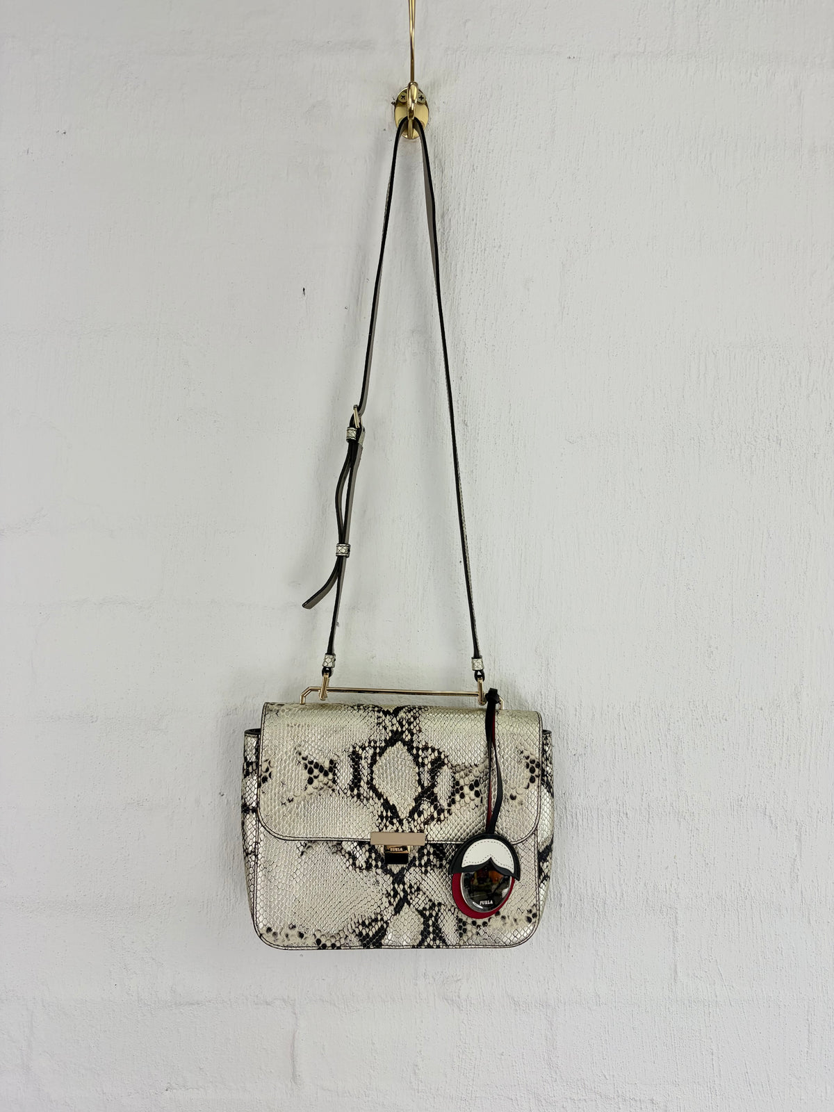 A small snakeskin-patterned handbag with a black shoulder strap and a round black-and-white charm, hanging against a white painted brick wall.