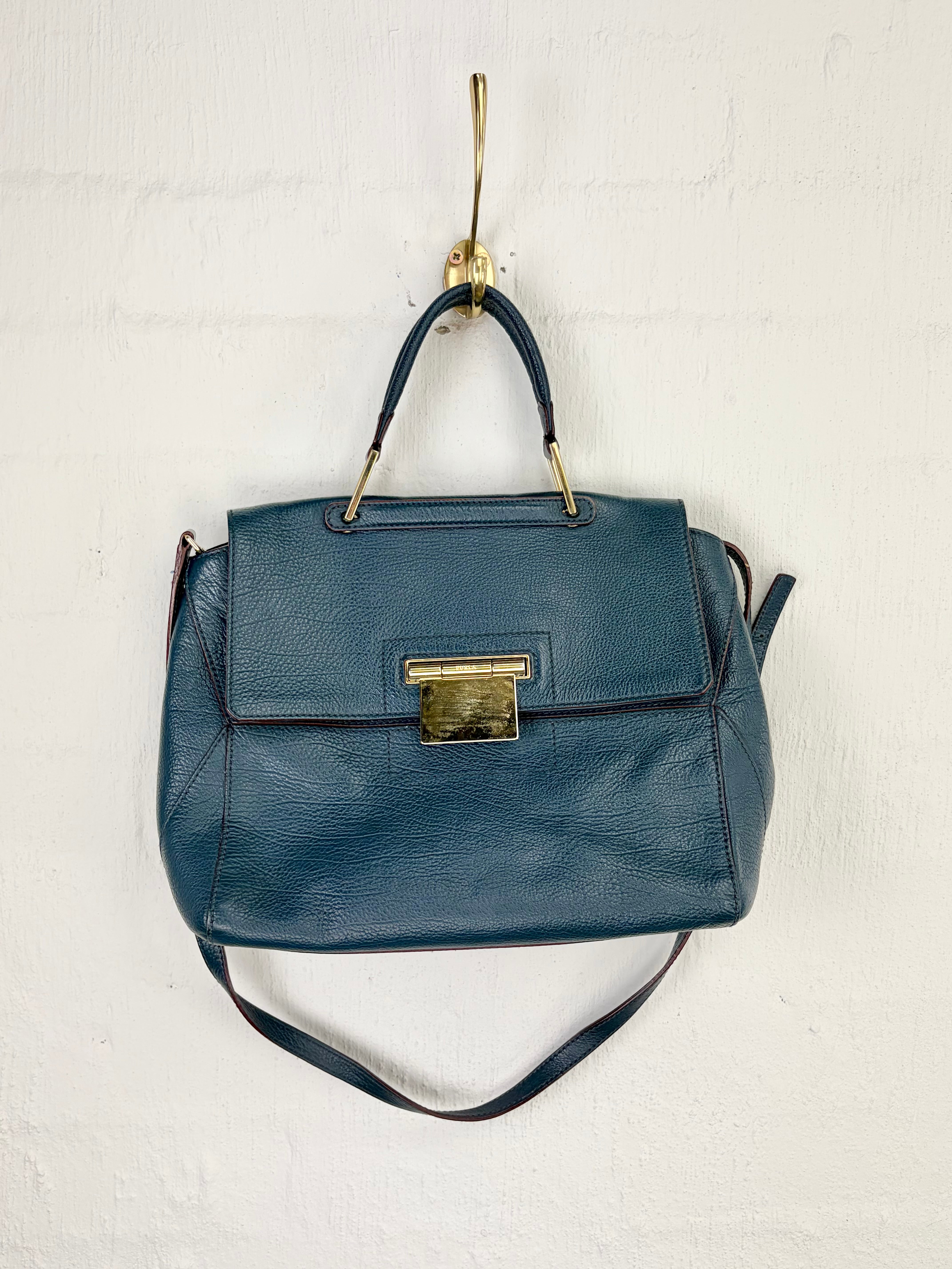 A dark blue pebbled leather handbag with gold-tone hardware, hanging from a gold wall hook against a white painted brick wall.