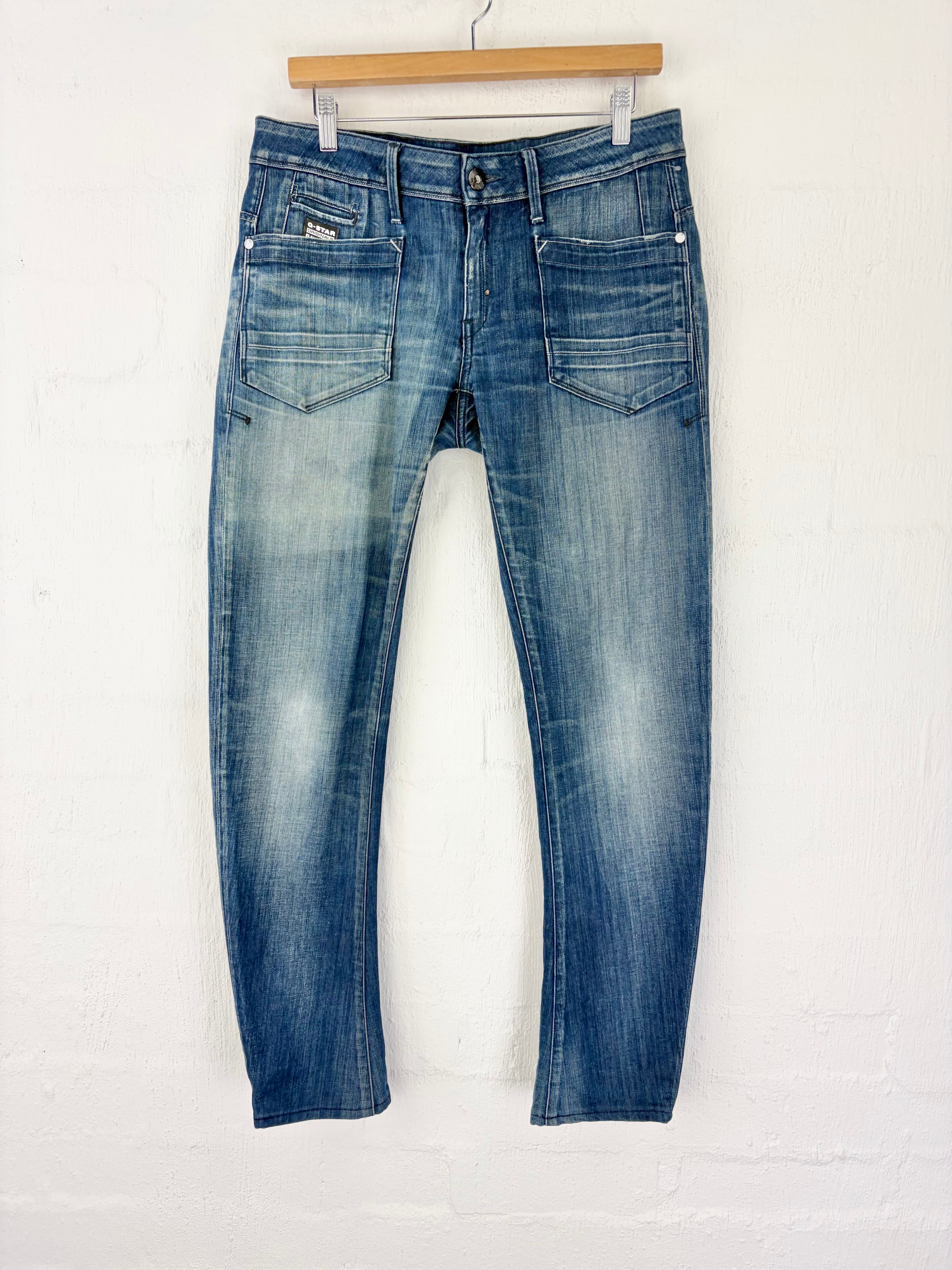 A pair of medium-wash blue denim jeans with a faded wash, hanging from a wooden hanger against a white painted brick wall.