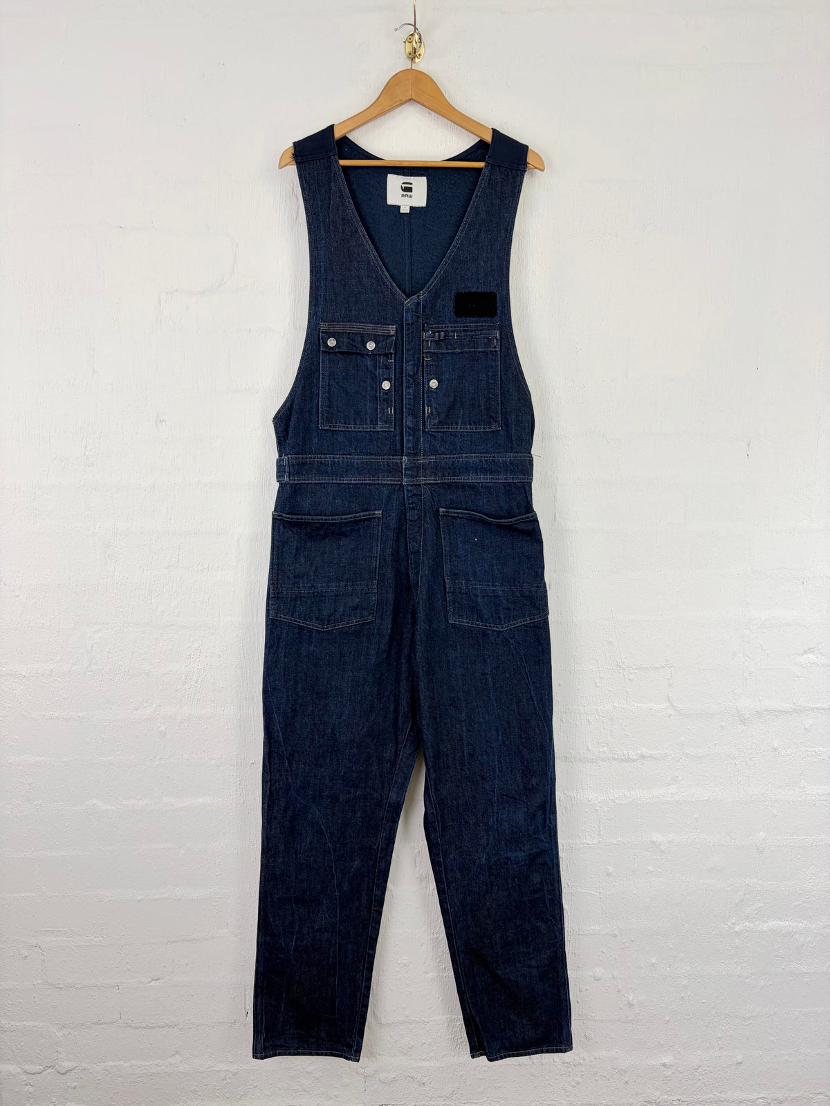 A dark indigo denim bib overalls hanging on a wooden hanger against a white painted brick wall. The overalls feature a V-neck, buttoned bib front, chest pockets with silver rivets, and large front cargo pockets.
