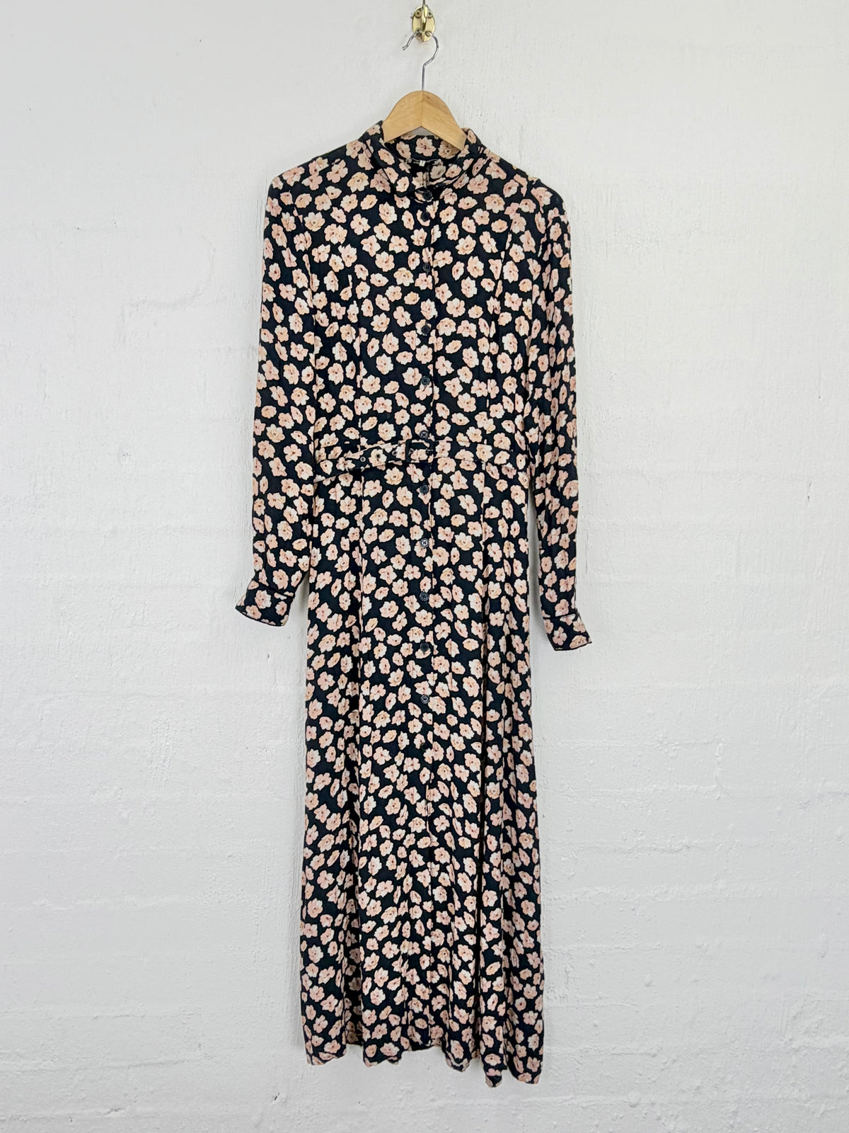 A long-sleeve midi dress with a high collar and an allover small floral print, hanging on a wooden hanger against a white painted brick wall.