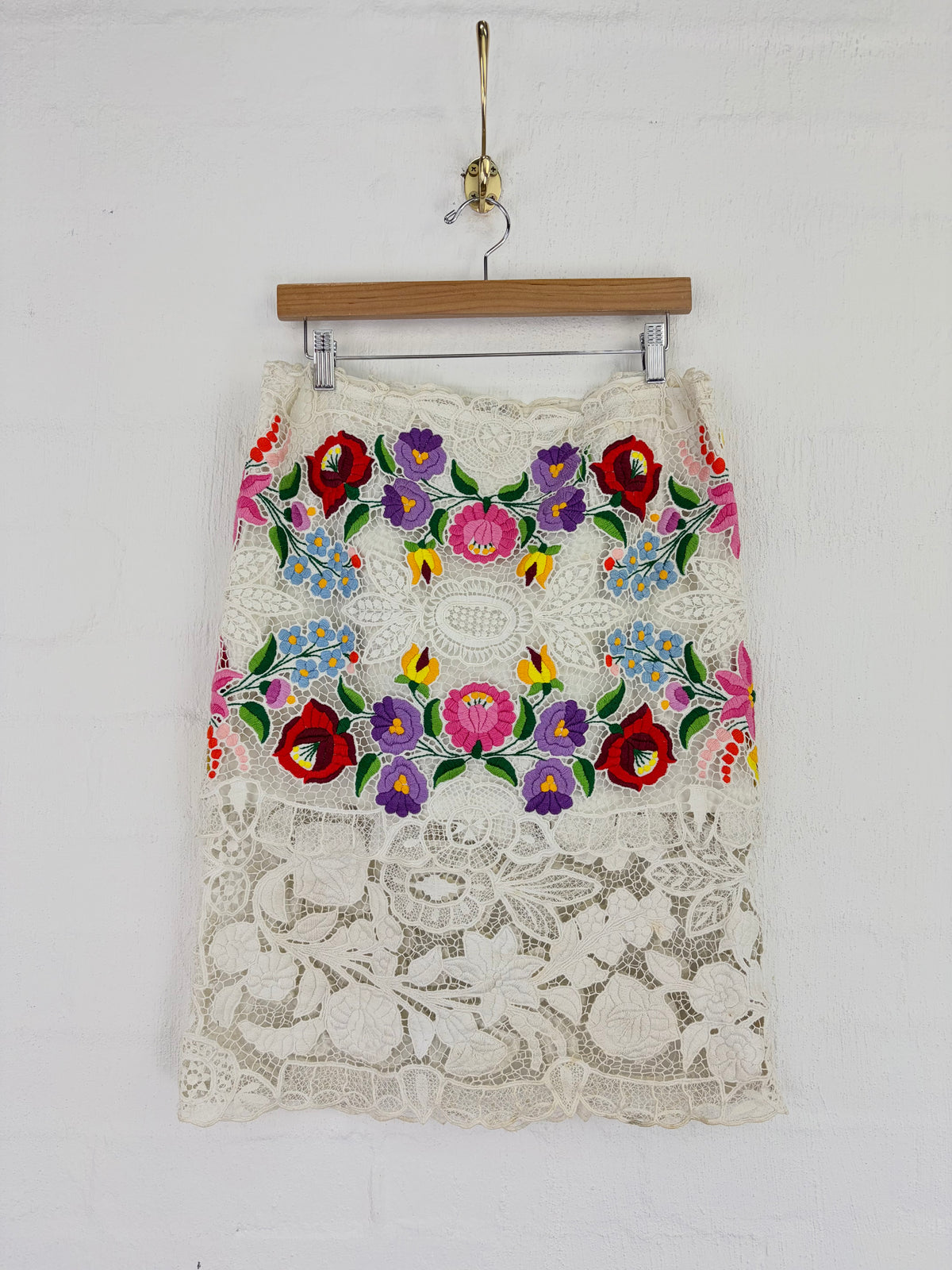 An off-white lace skirt with colorful floral embroidery, hanging from a wooden hanger against a white painted brick wall.