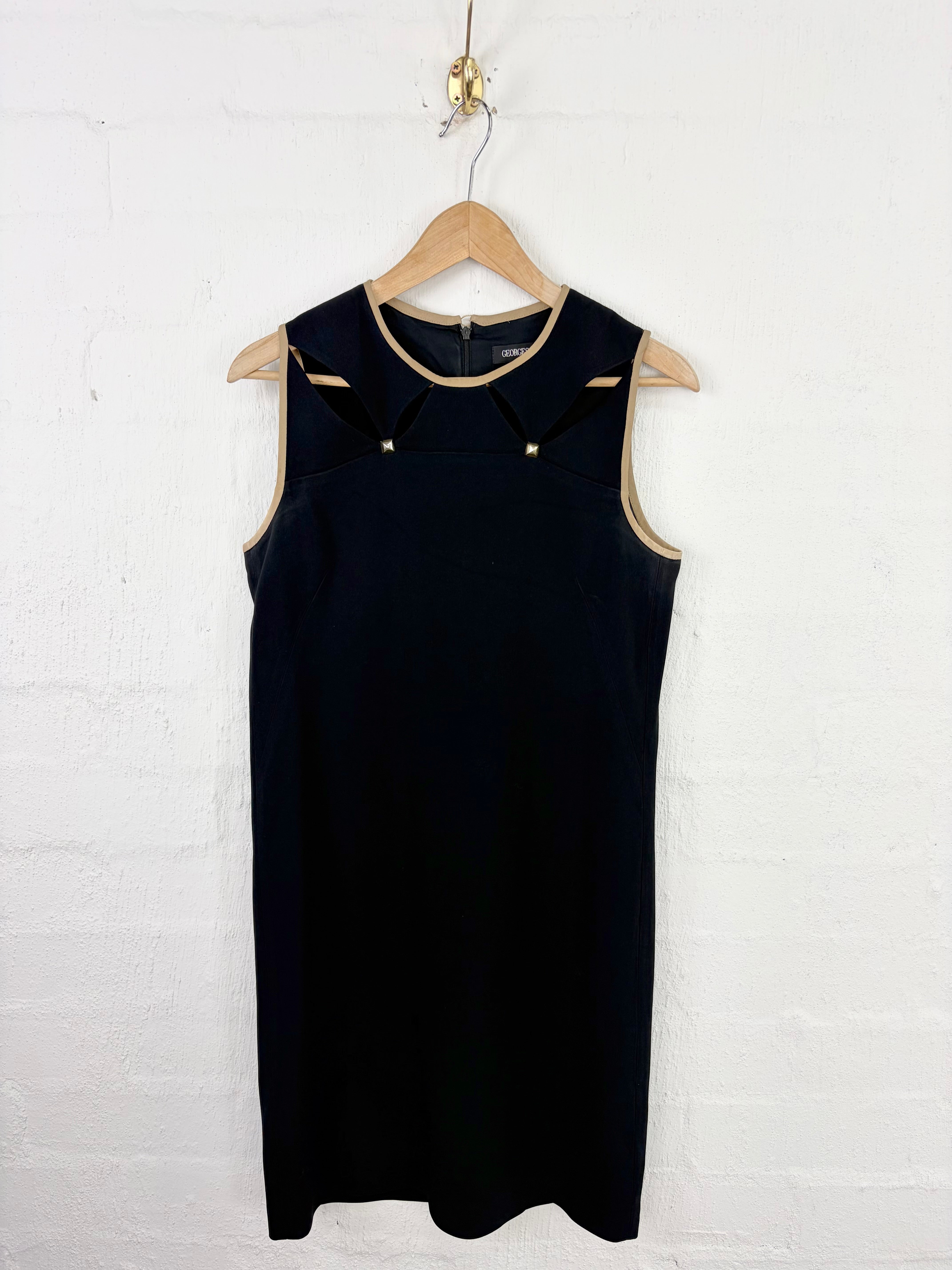 A sleeveless black dress with beige trim and small gold-tone decorative details, hanging on a wooden hanger against a white painted brick wall.