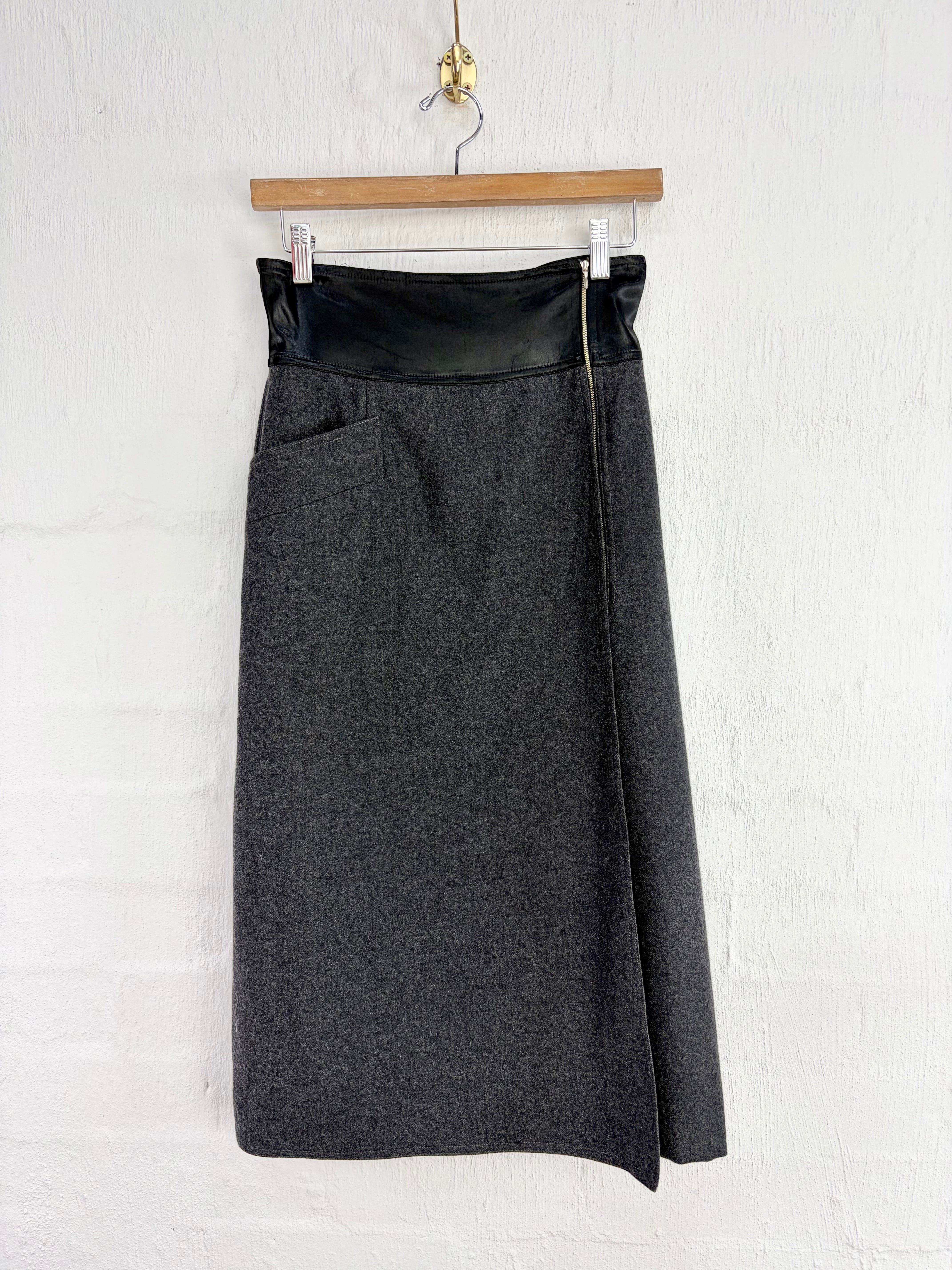 A dark gray textured wool skirt with a black faux-leather waistband and a side zipper detail, hanging from a wooden hanger against a white brick wall.
