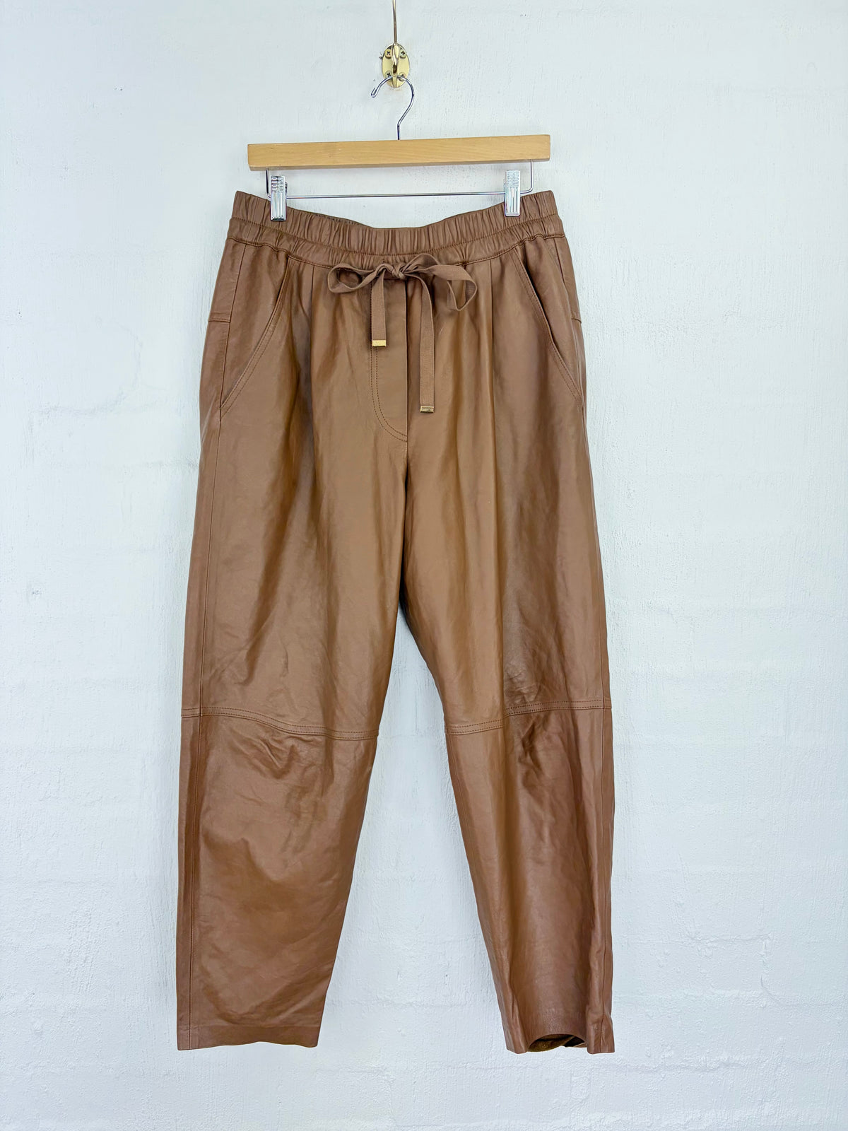 A pair of brown faux leather cropped pants with an elastic drawstring waist, hanging from a wooden hanger against a white brick wall.