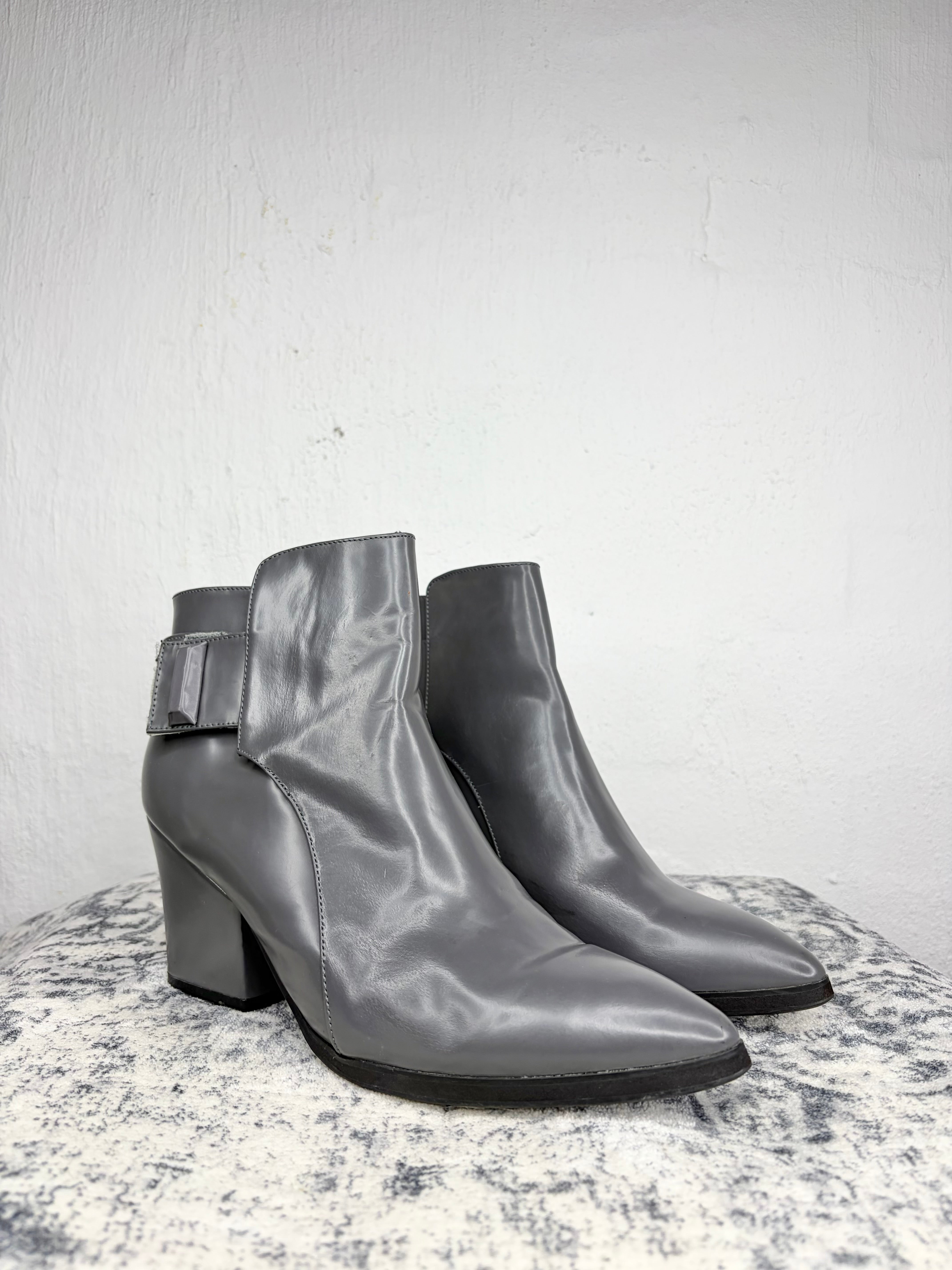 A pair of women's gray leather ankle boots with a block heel and a decorative buckle strap on the outer side, displayed on a textured stone surface against a white wall.