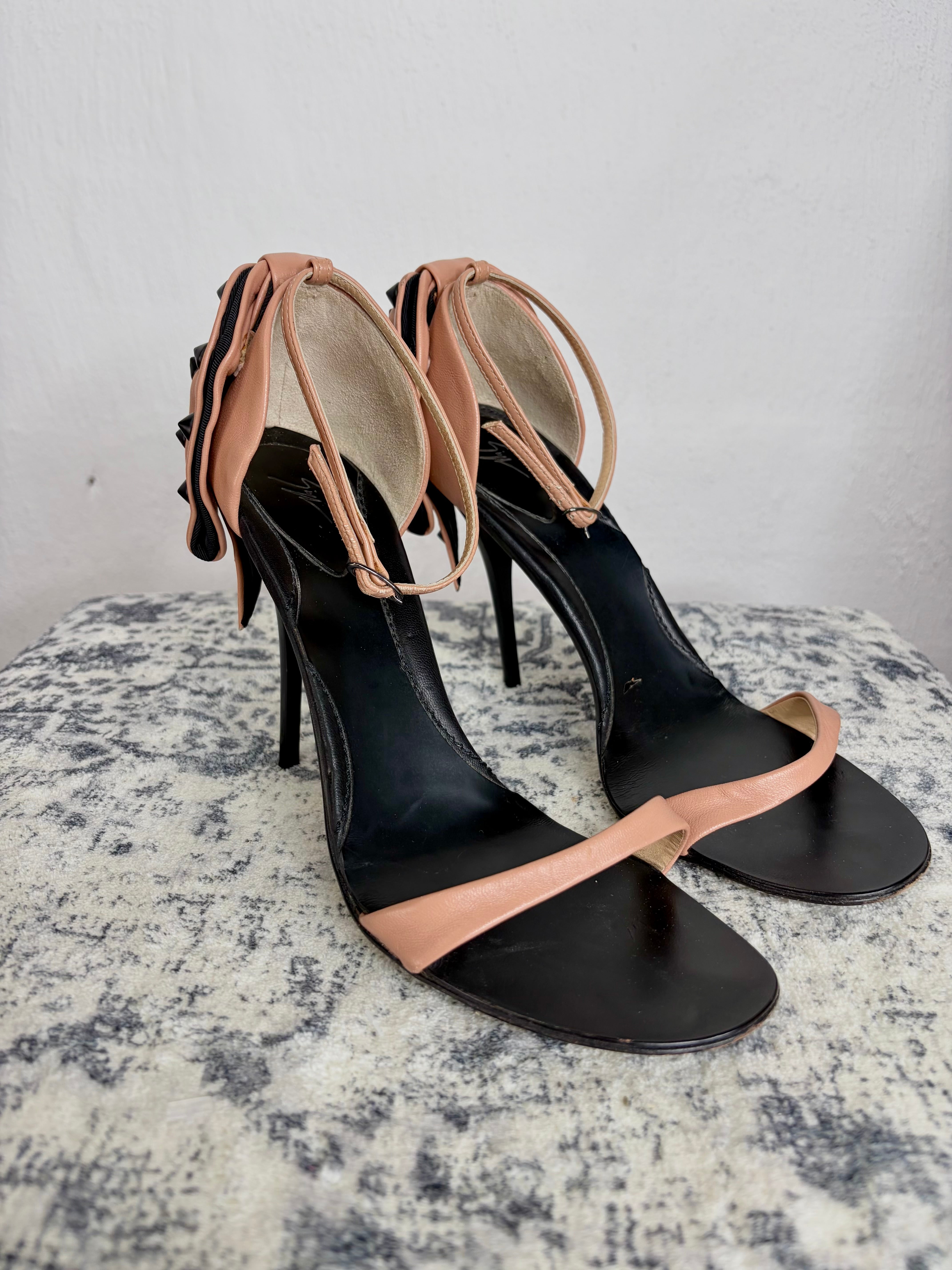 A pair of black leather high-heeled sandals with nude beige straps, displayed on a textured gray surface against a light gray wall.