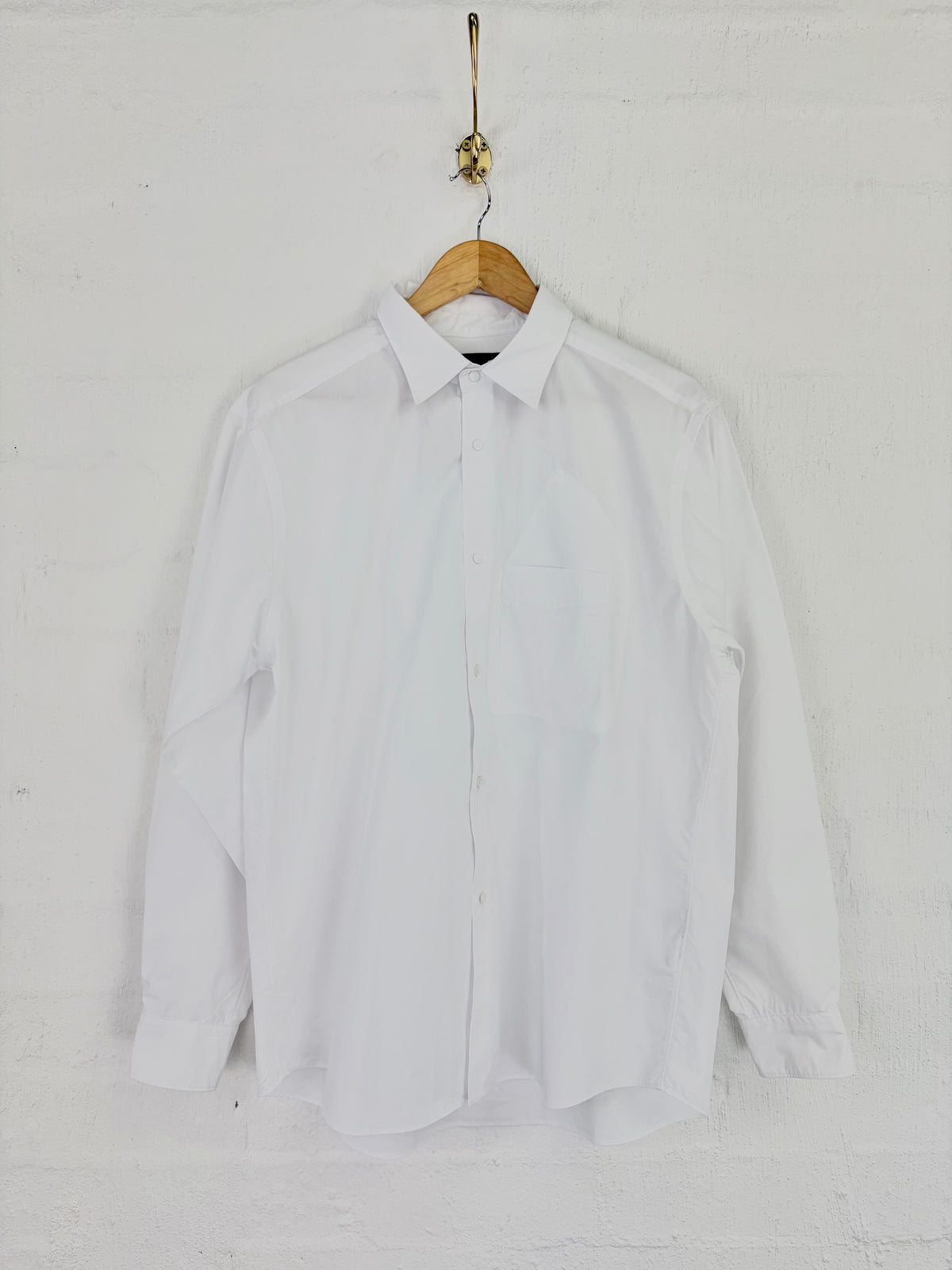 A white long-sleeve button-up shirt hanging on a wooden hanger against a white painted brick wall.