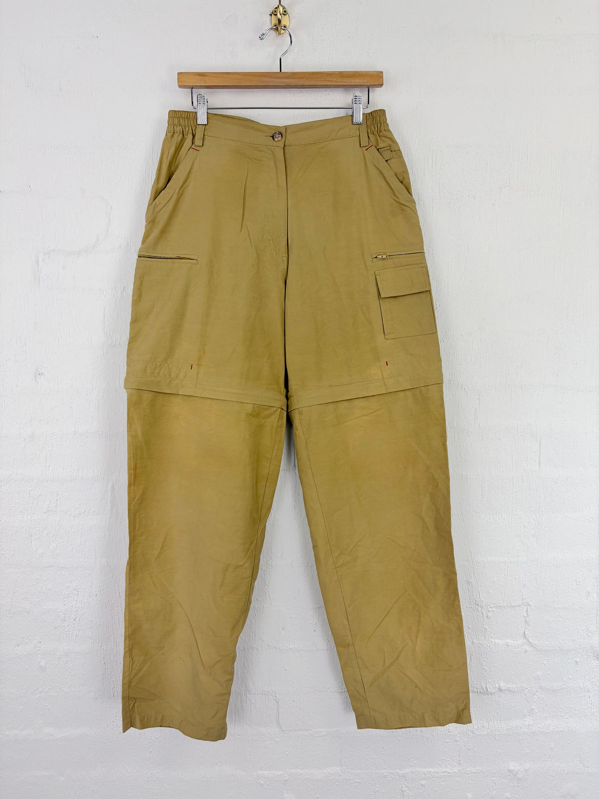 A pair of olive green convertible cargo pants hanging from a wooden hanger against a white painted brick wall.