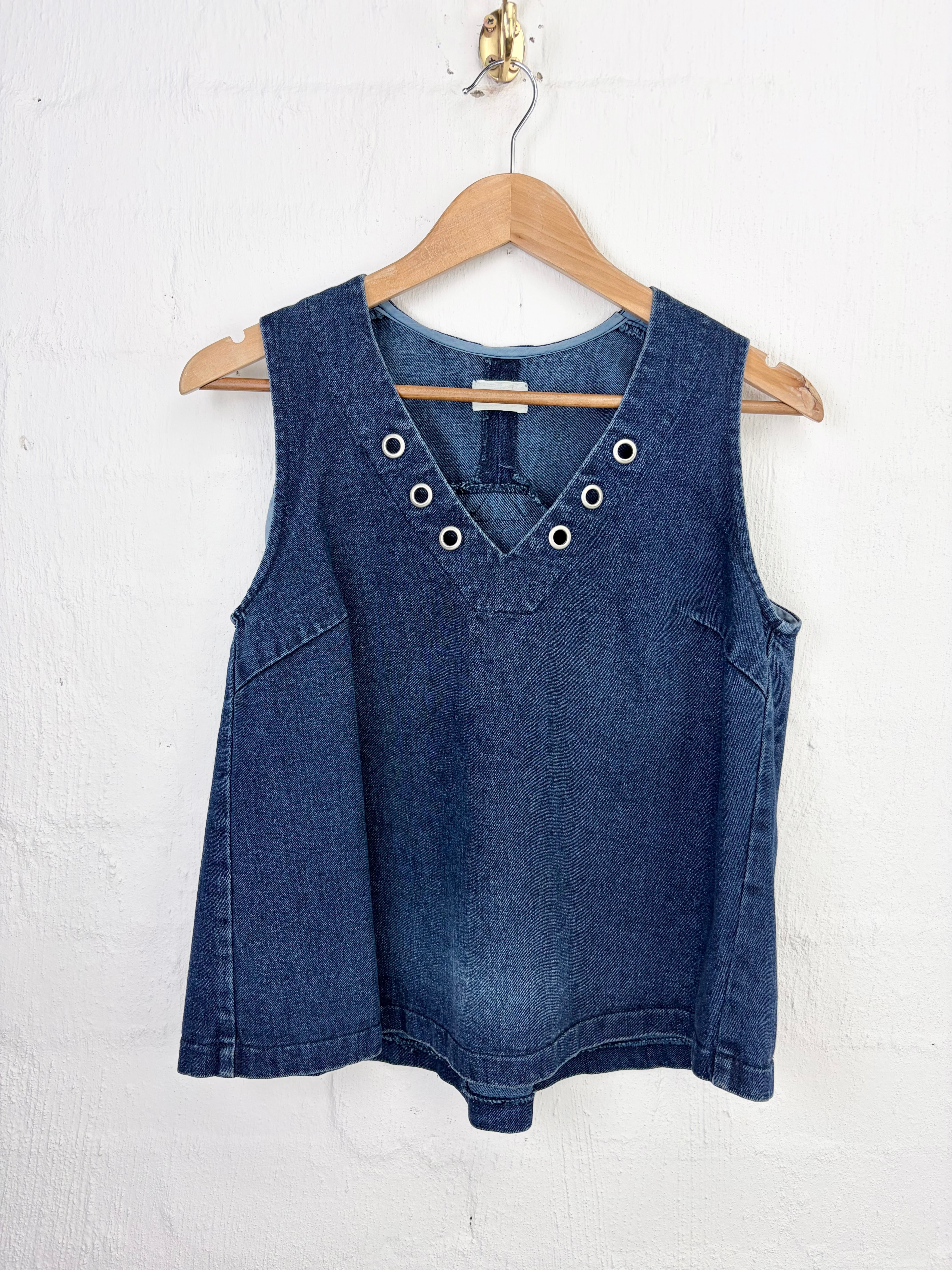 A sleeveless dark blue denim vest with decorative metal eyelets on the neckline, hanging on a wooden hanger against a white textured wall.