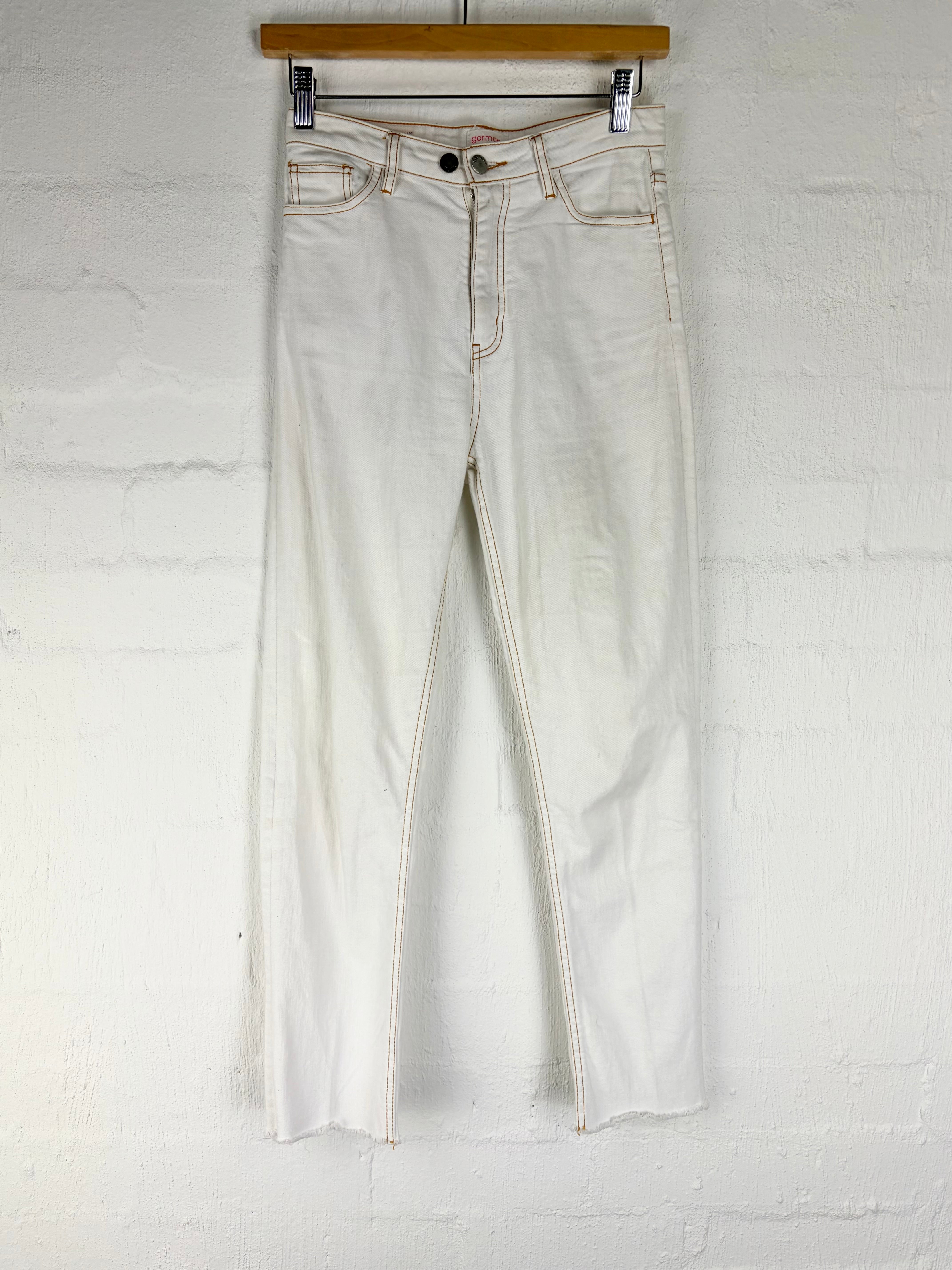 A pair of light off-white or cream-colored denim jeans hanging from a wooden hanger against a white painted brick wall.