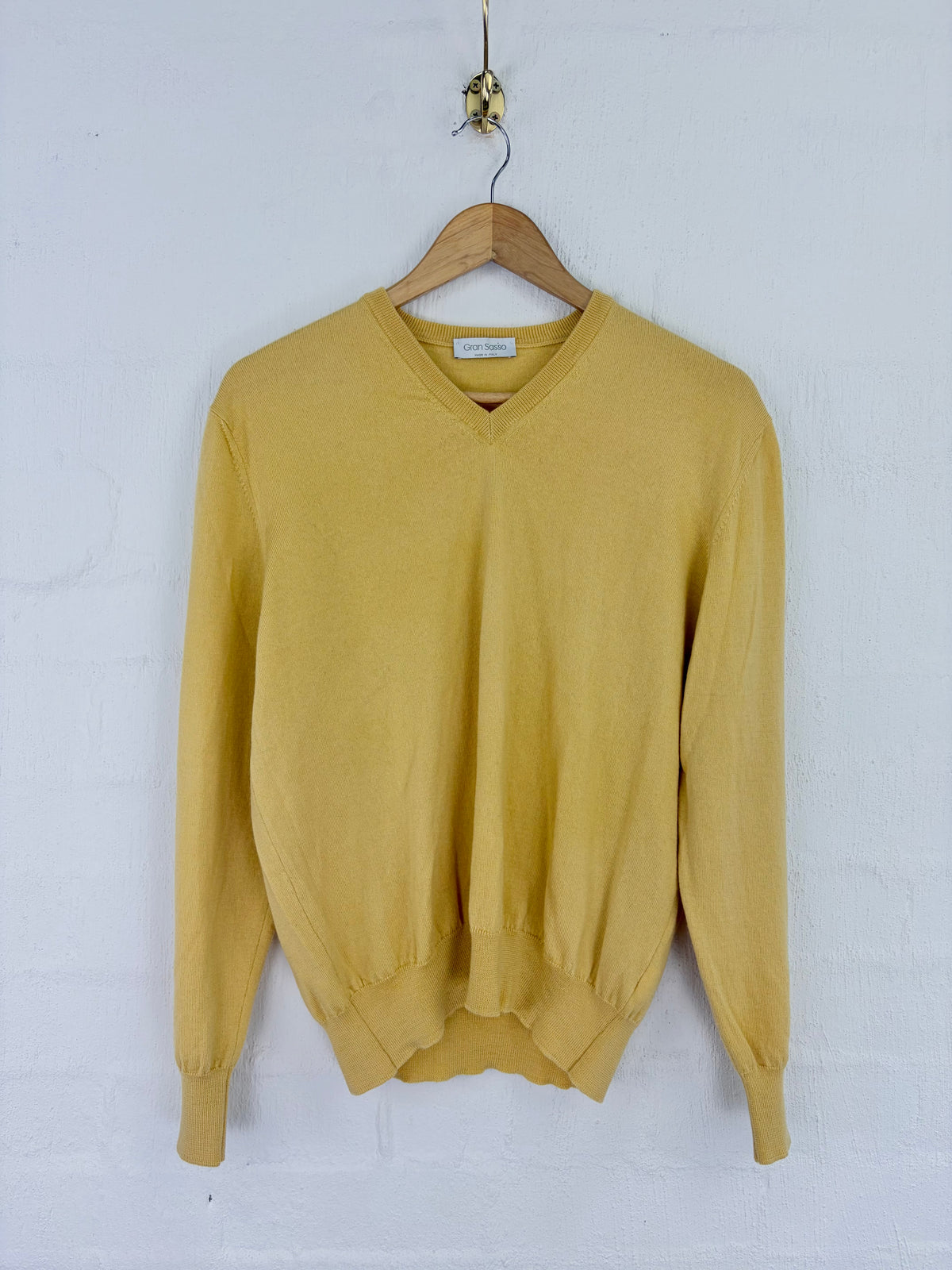A mustard yellow V-neck knit sweater hanging on a wooden hanger against a white painted brick wall.
