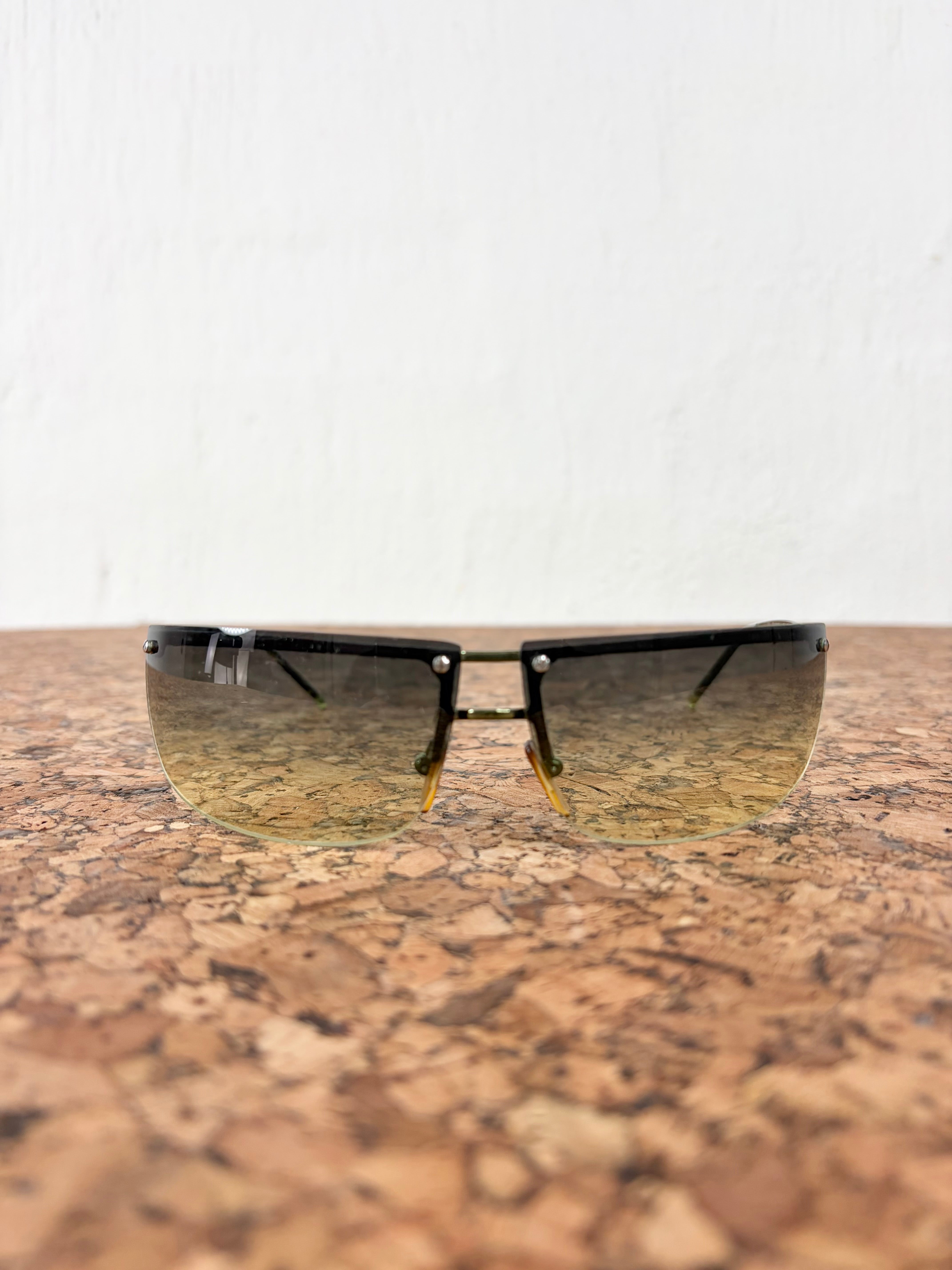 A pair of rimless shield-style sunglasses with a black frame and gradient tinted lenses, resting on a textured cork surface against a white wall.