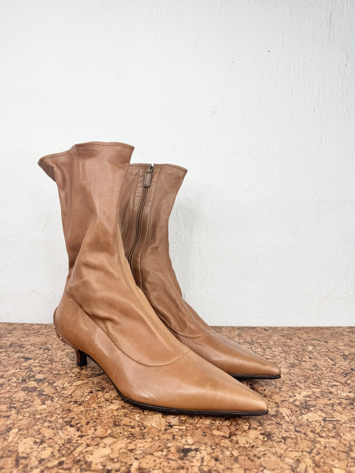 A pair of tan leather ankle boots with pointed toes and low kitten heels, standing on a brown speckled surface against a white wall.