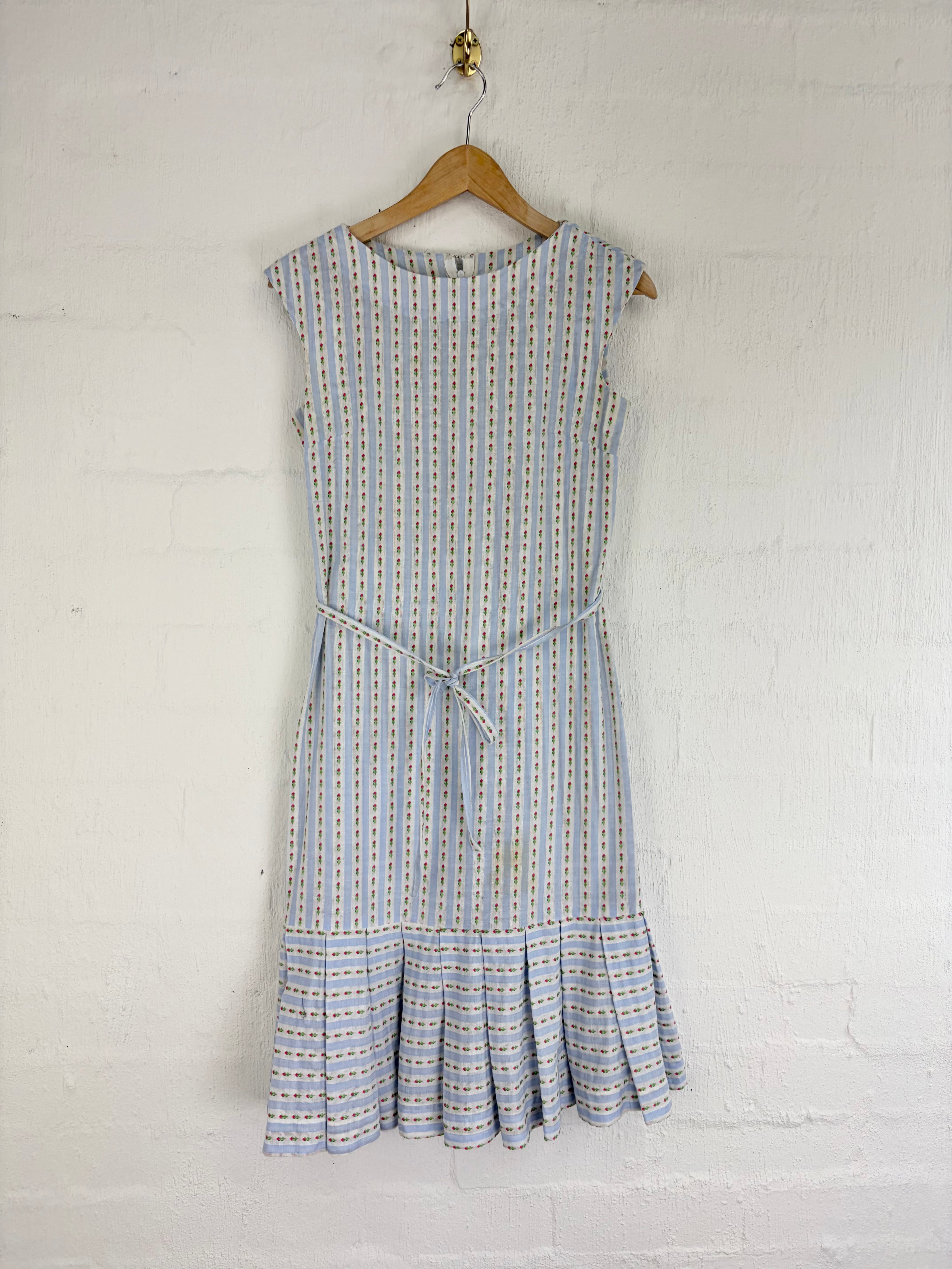 A sleeveless light blue and white vertically striped dress with a matching tie belt and ruffled hem, hanging on a wooden hanger against a white painted brick wall.