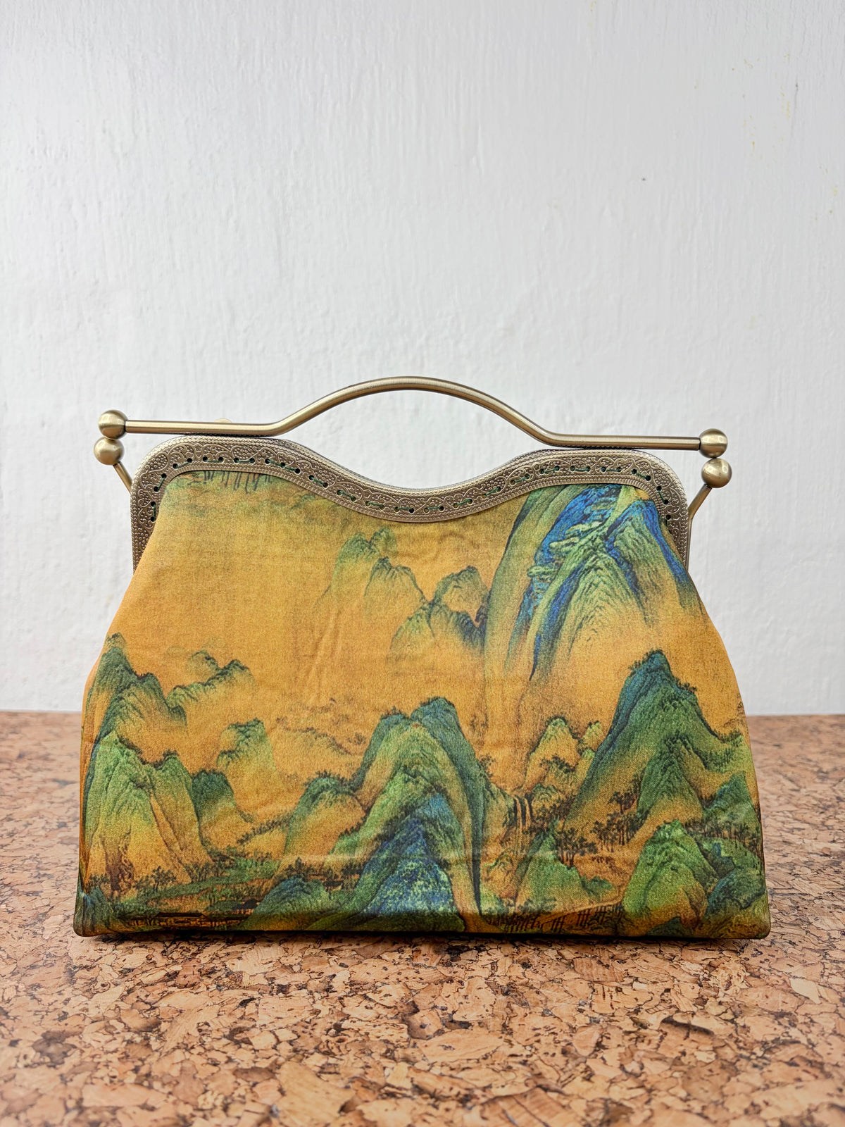 A vintage-style handbag with a gold-tone metal frame handle, featuring a printed fabric body with a landscape design of green and blue mountains on a golden-yellow background, resting on a cork surface against a white wall.