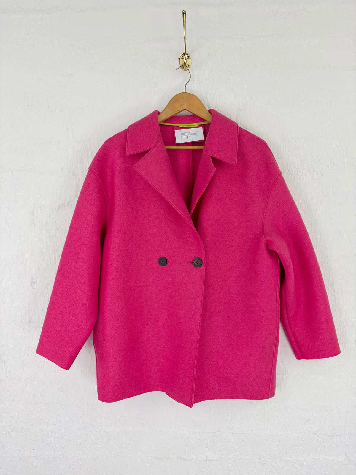 A bright pink double-breasted coat with notched lapels and two black buttons, hanging on a wooden hanger against a white painted brick wall.