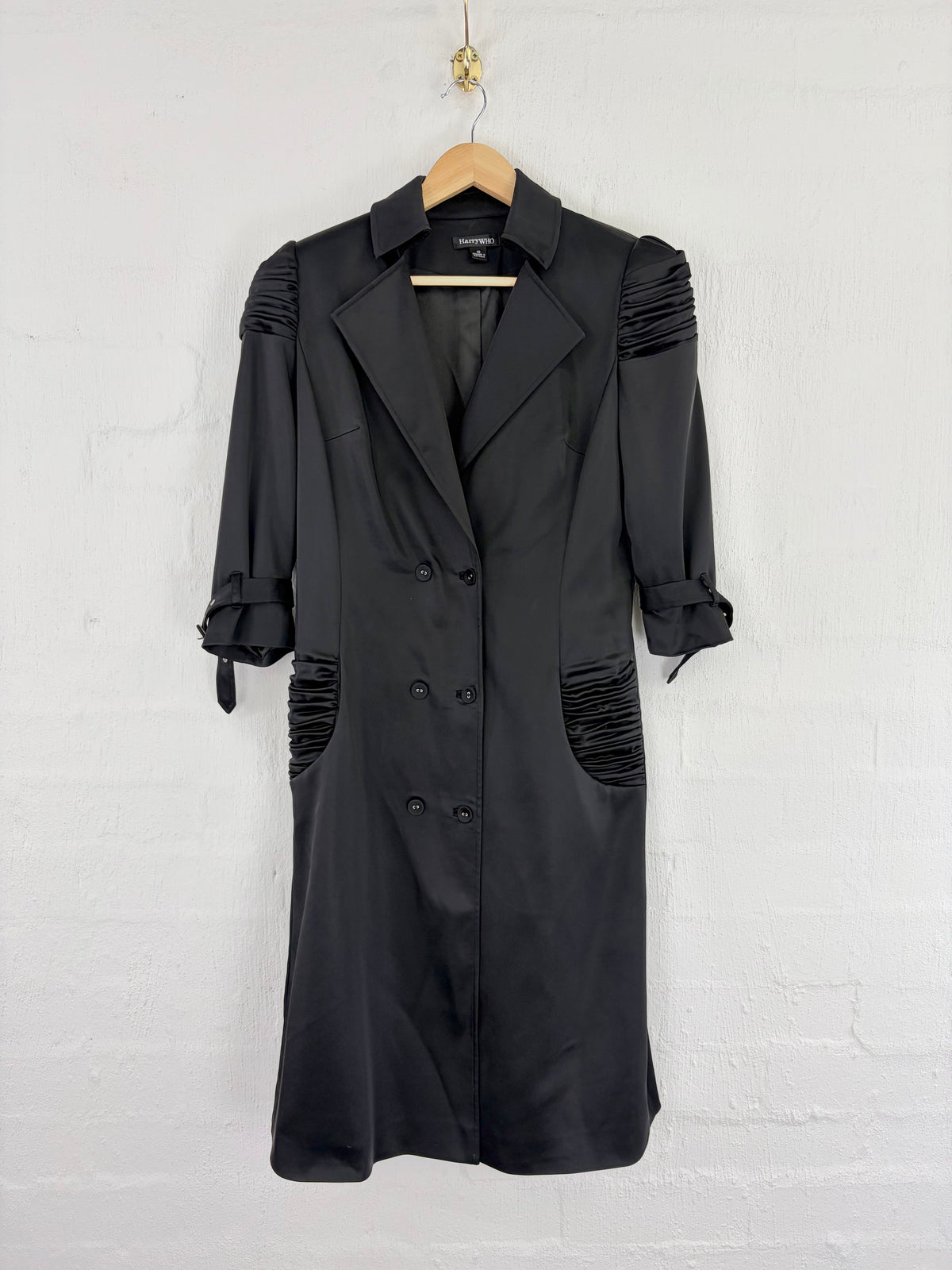A black double-breasted trench coat with ruched shoulder and hem details, hanging on a wooden hanger against a white painted brick wall.