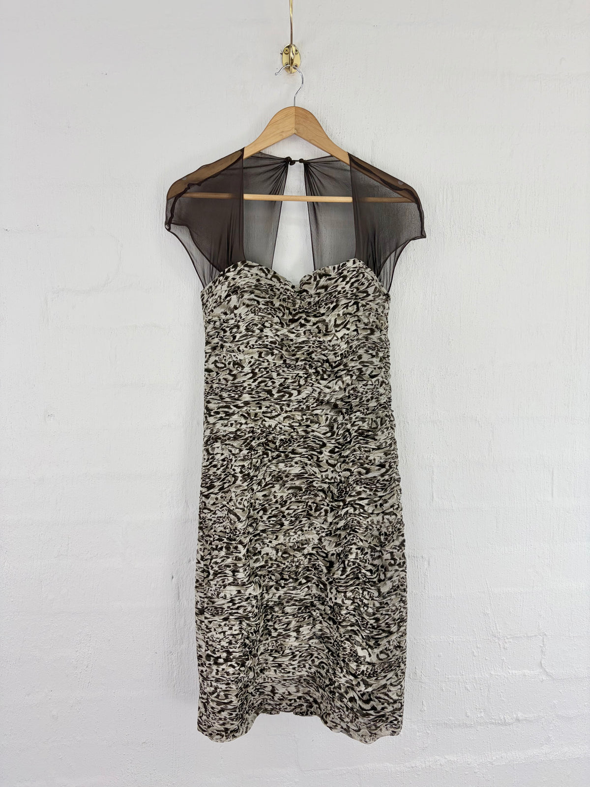 A sleeveless sheath dress with a black sheer mesh yoke and a textured black, gray, and white patterned skirt, hanging on a wooden hanger against a white brick wall.