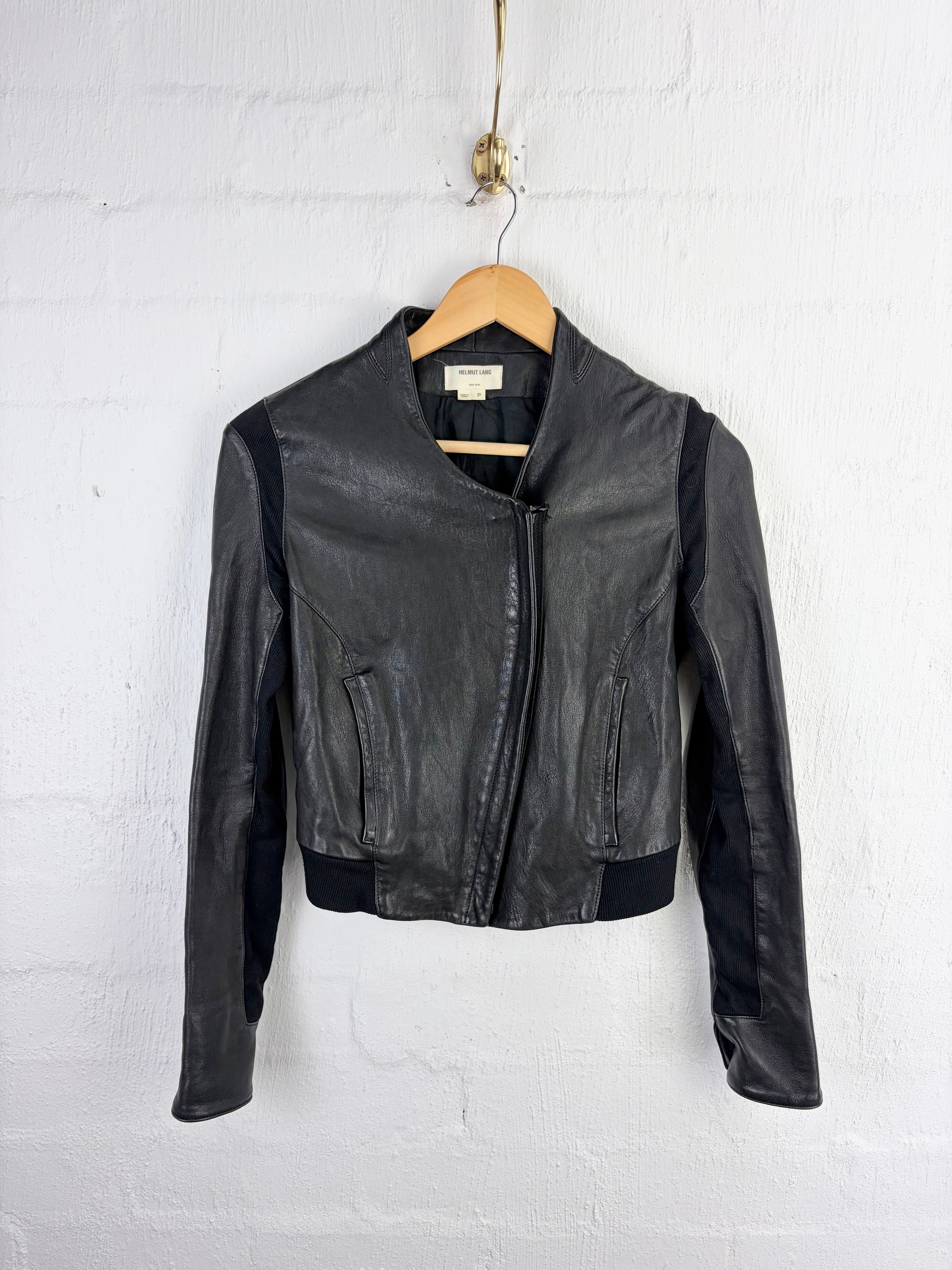 A black leather jacket with contrasting black fabric side panels, hanging on a wooden hanger against a white painted brick wall.