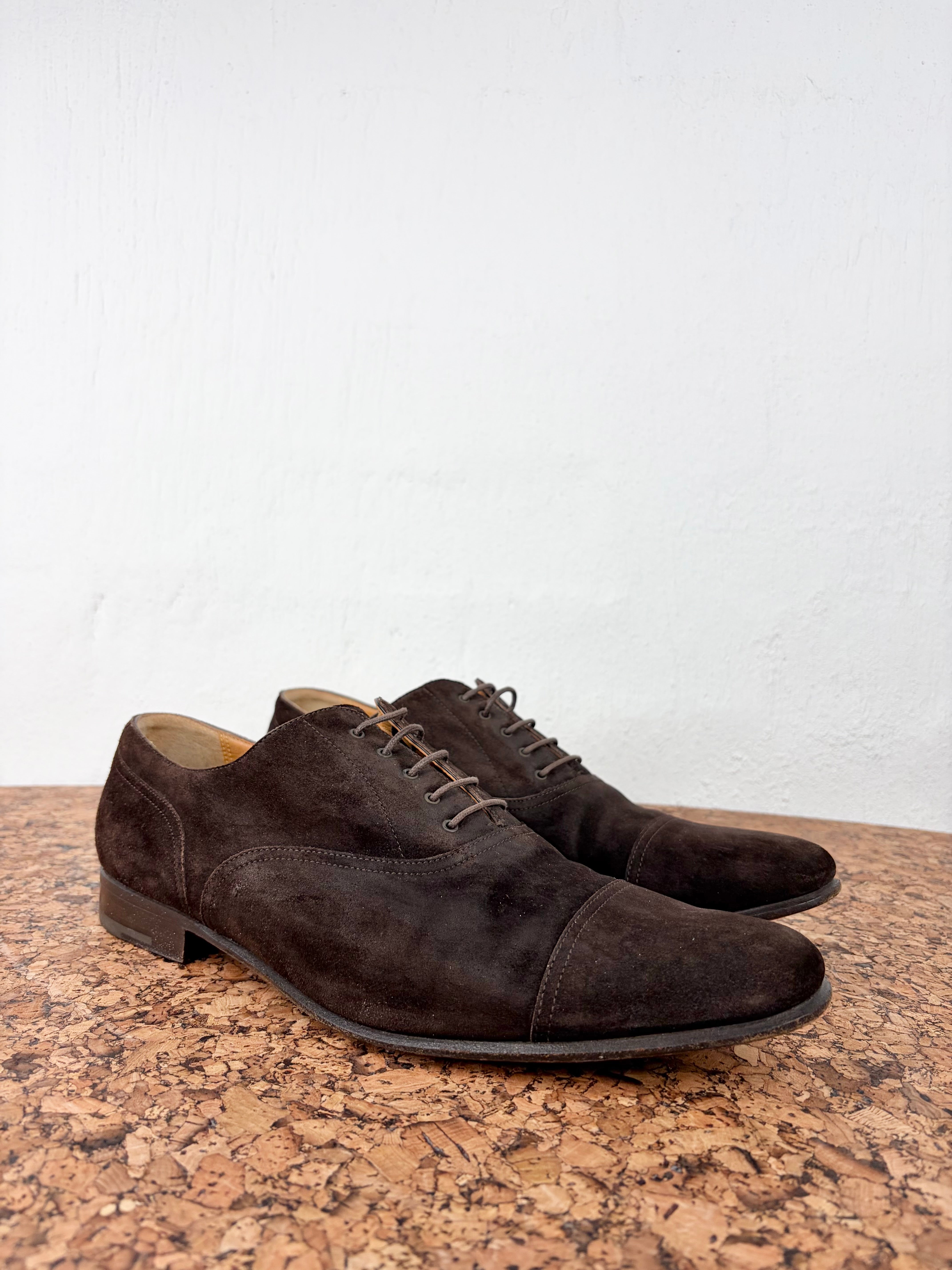 A pair of dark brown suede cap-toe oxford shoes resting on a textured cork surface against a plain white wall.