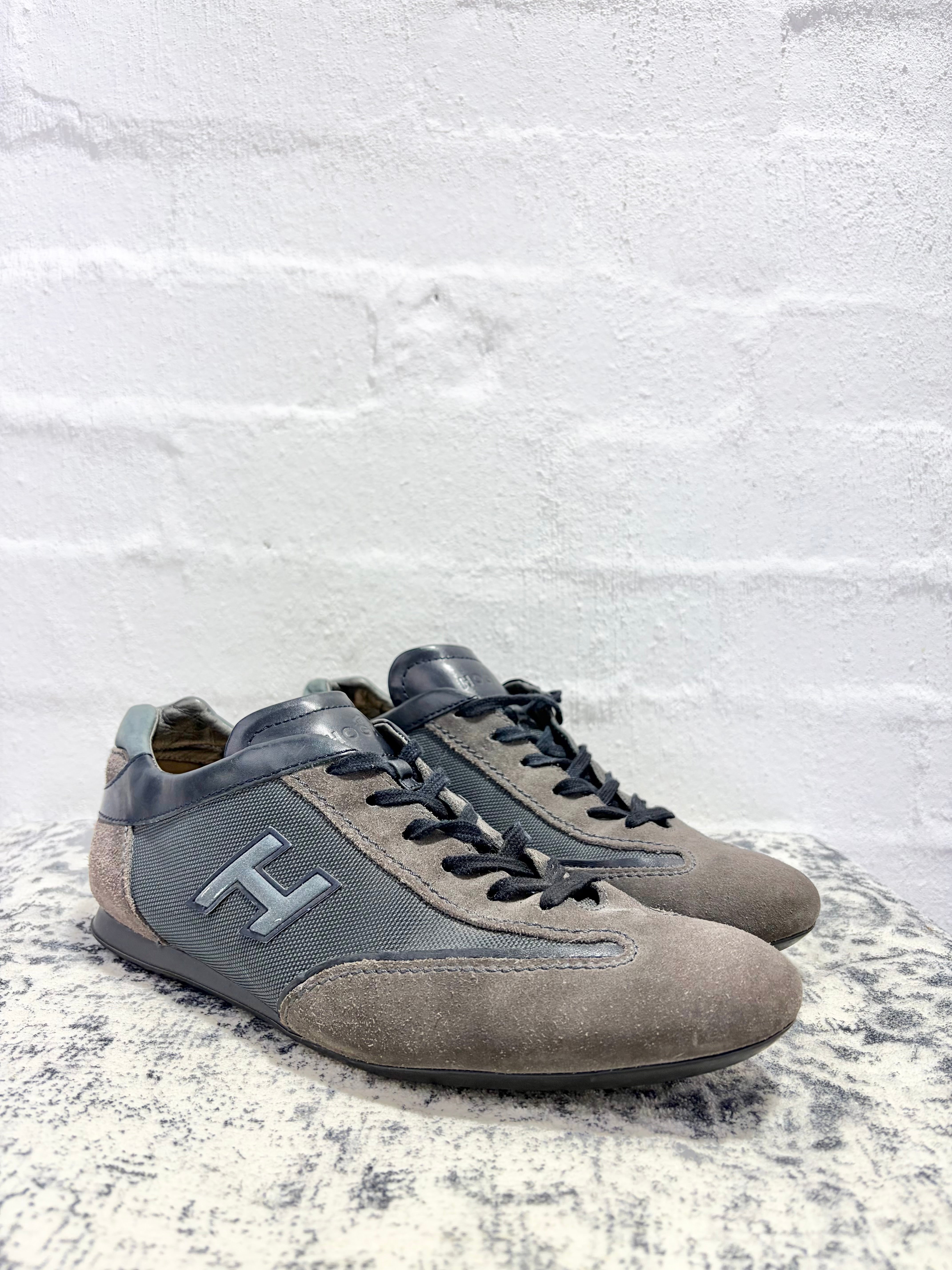 A pair of grey suede and mesh low-top sneakers with black laces and a prominent grey 'H' logo on the side, displayed on a textured grey stone surface against a white brick wall.