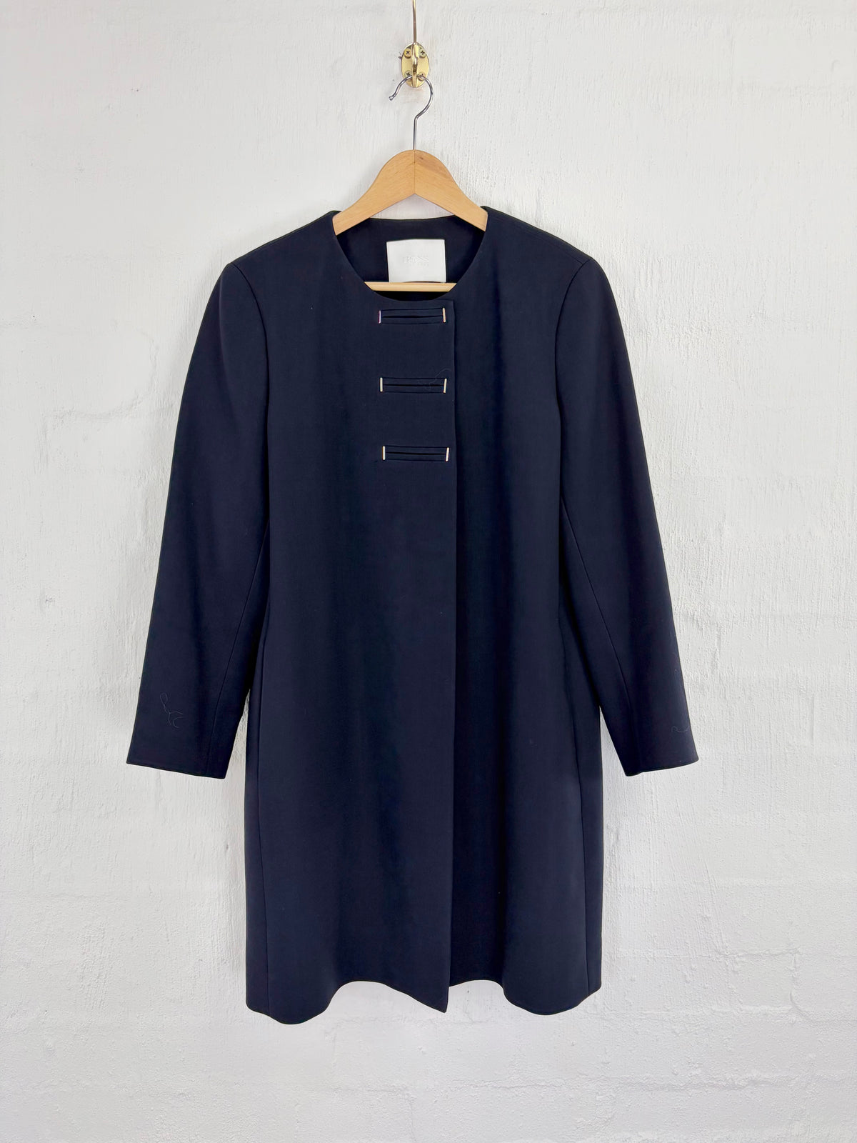 A dark navy blue long-sleeve tunic dress with three decorative button details on the front, hanging on a wooden hanger against a white painted brick wall.