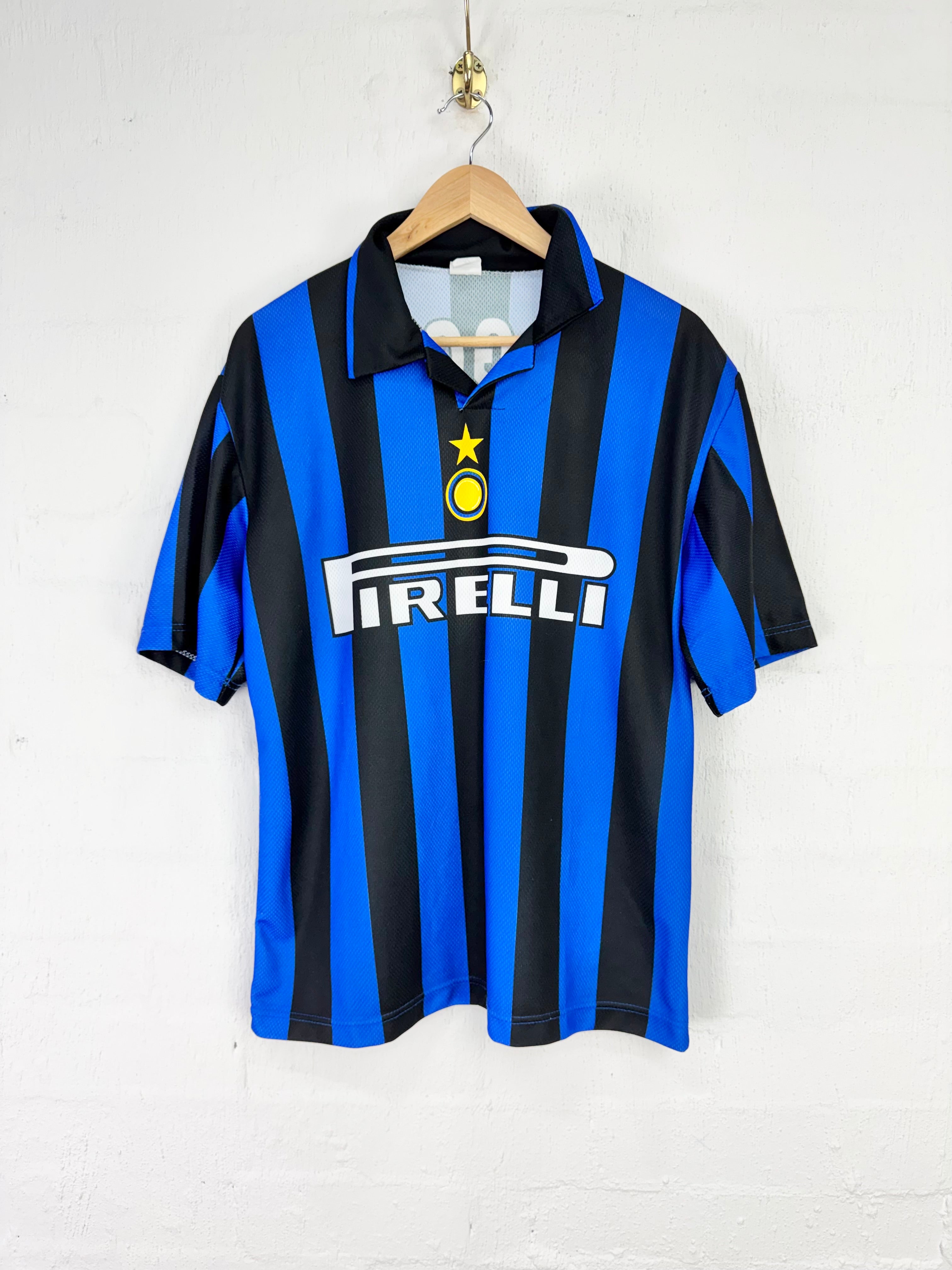 A vintage Inter Milan soccer jersey with blue and black vertical stripes, featuring the Pirelli sponsor logo on the chest, hanging on a wooden hanger against a white brick wall.
